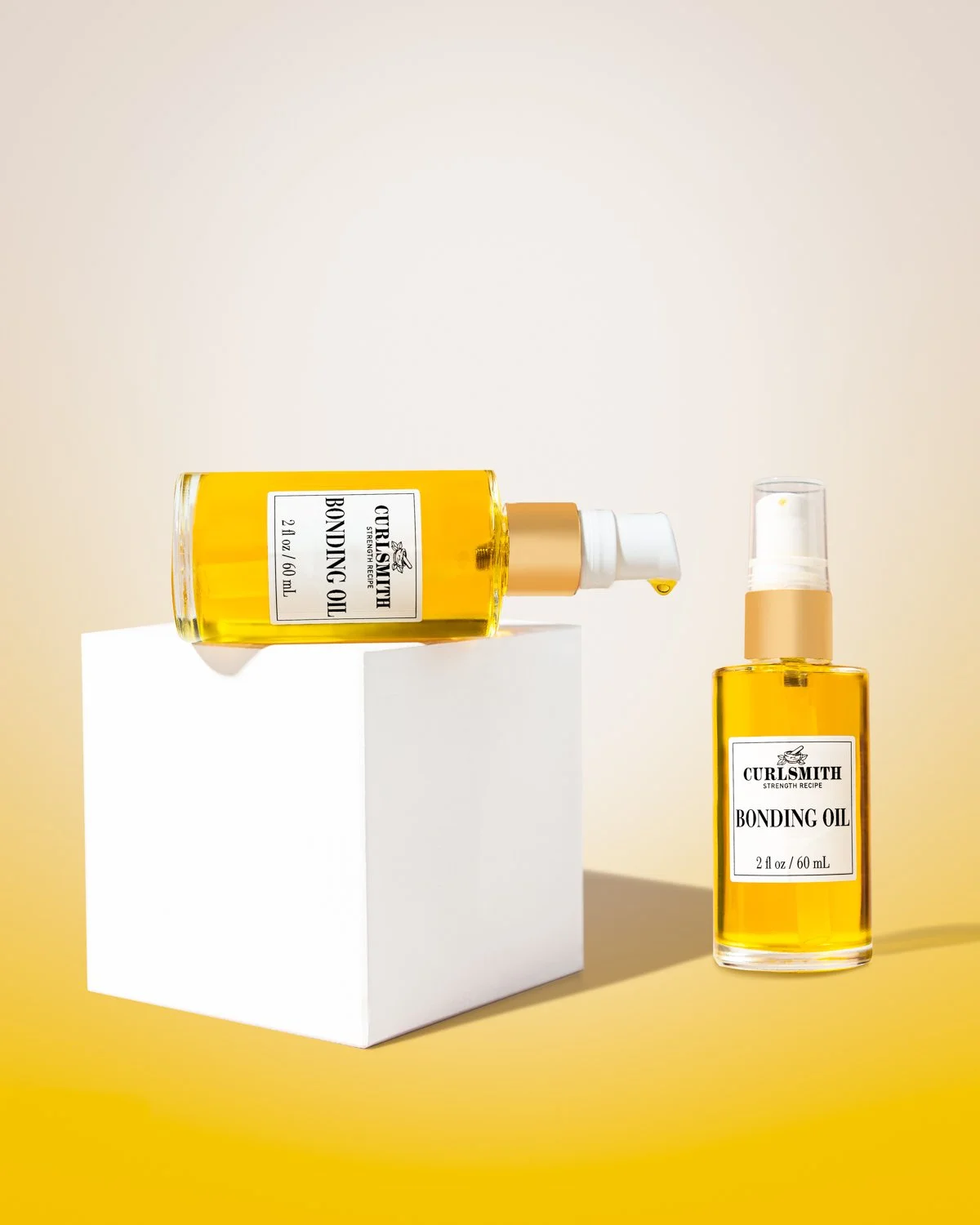 Two bottles of curlsmith bonding oil, one lying on a white block with a drop coming out, and the other standing upright, set against a yellow background.