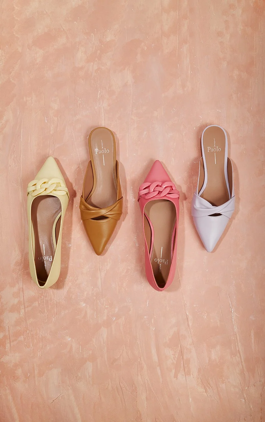 Four pointed-toe flats in yellow, brown, pink, and white, each with braided straps on the upper, placed on a pink tile floor.