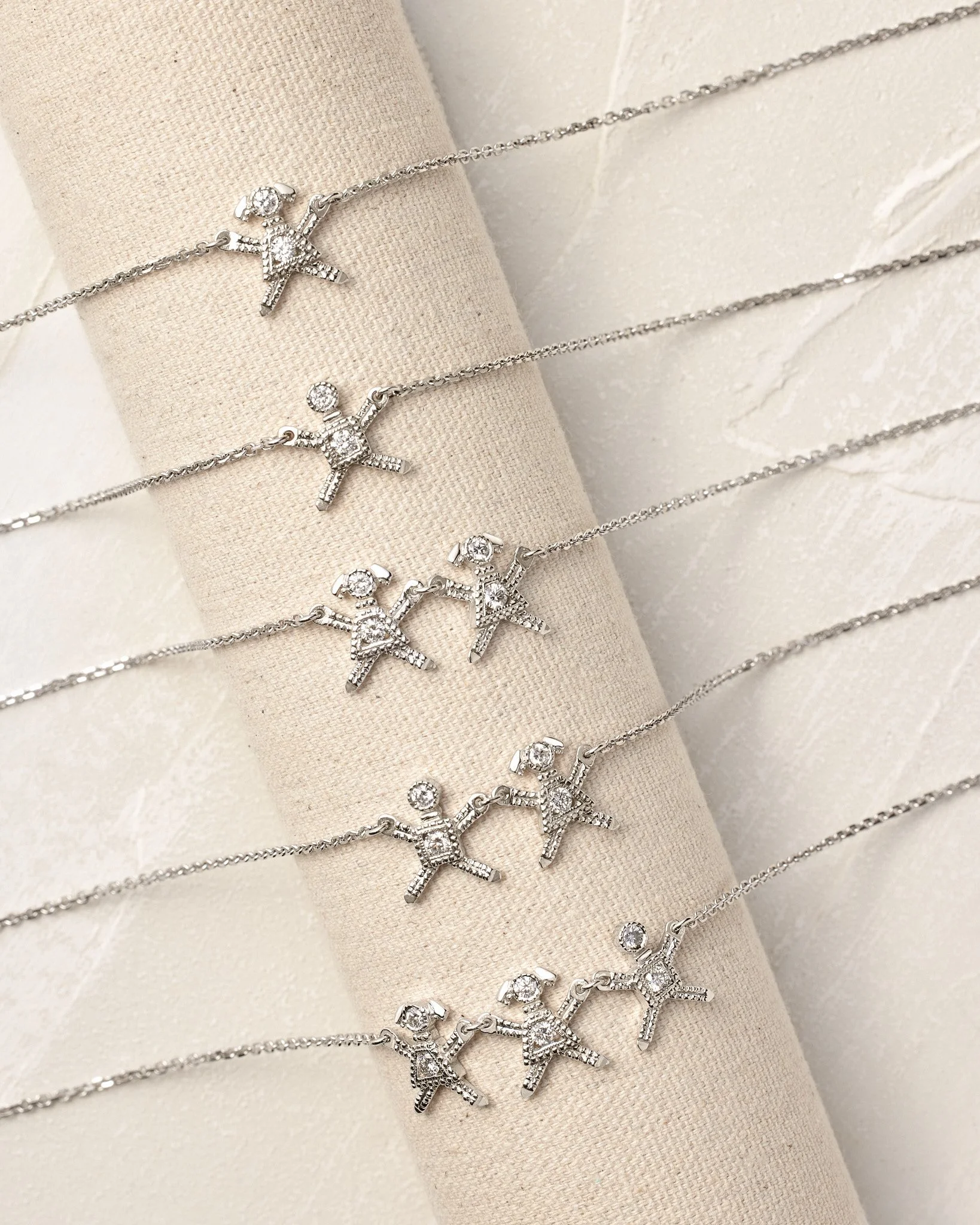 Multiple silver necklaces with child or doll-shaped pendants, each adorned with small stones, displayed on a beige fabric jewelry holder.