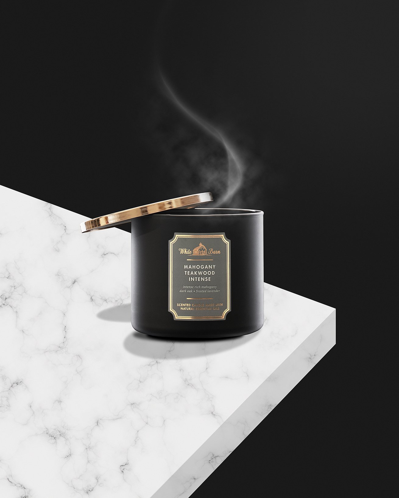 A black scented candle labeled 'Mahogany Teakwood Intense' with smoke rising from it, placed on a white marble surface against a black background.