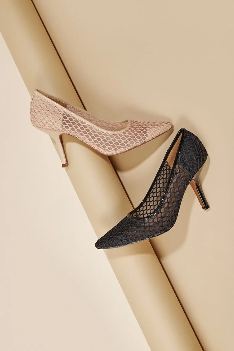 Two high-heeled shoes with fishnet pattern, one beige and one black, placed on a beige surface.