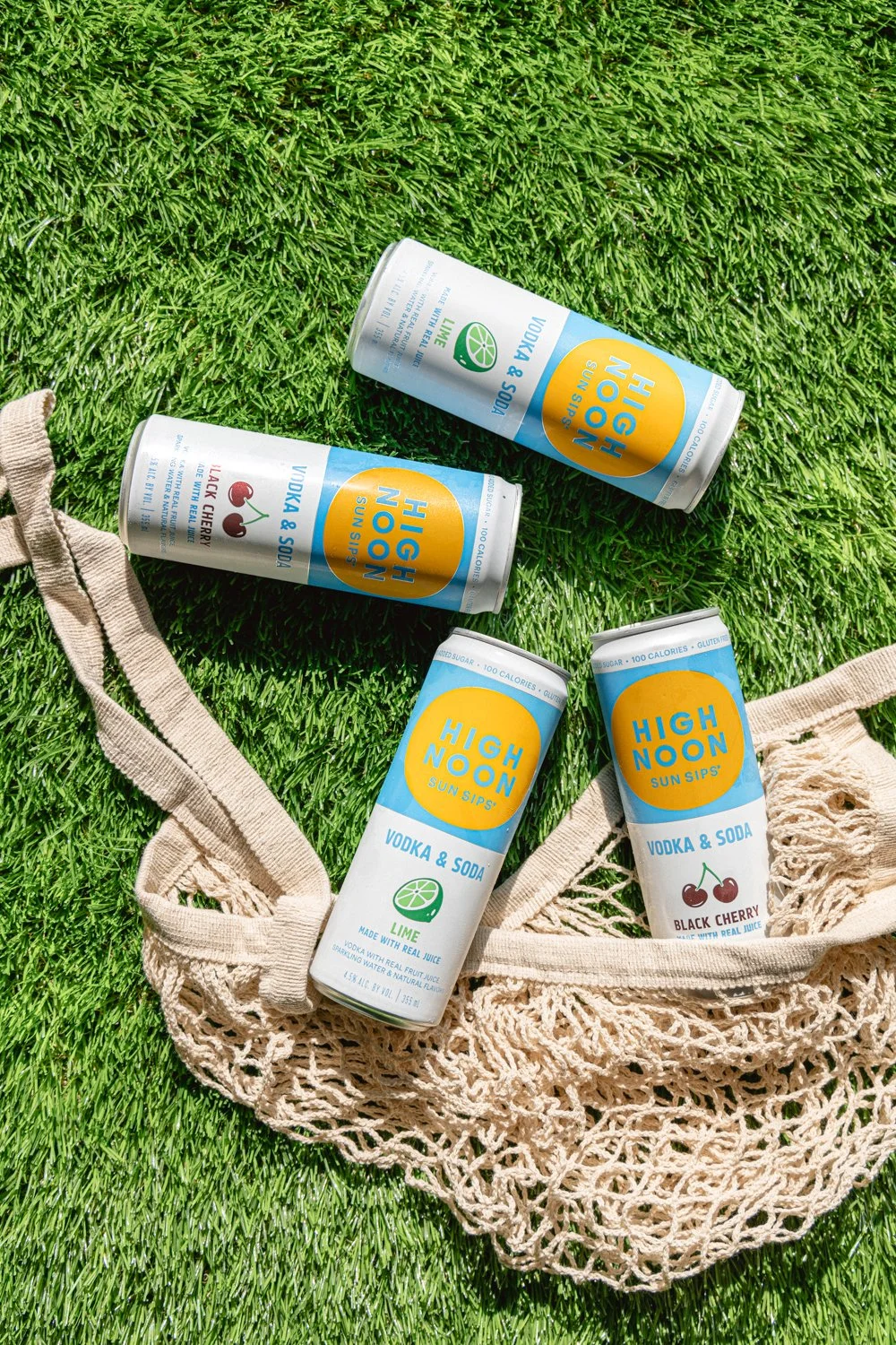 Four cans of High Noon vodka and soda, flavored with lime and black cherry, are placed on a grassy surface with a beige reusable bag nearby.