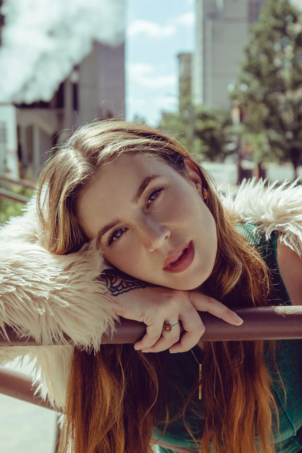 A woman lying with a faux fur coat outdoors on a sunny day, resting her head on her hand, with a cityscape background.