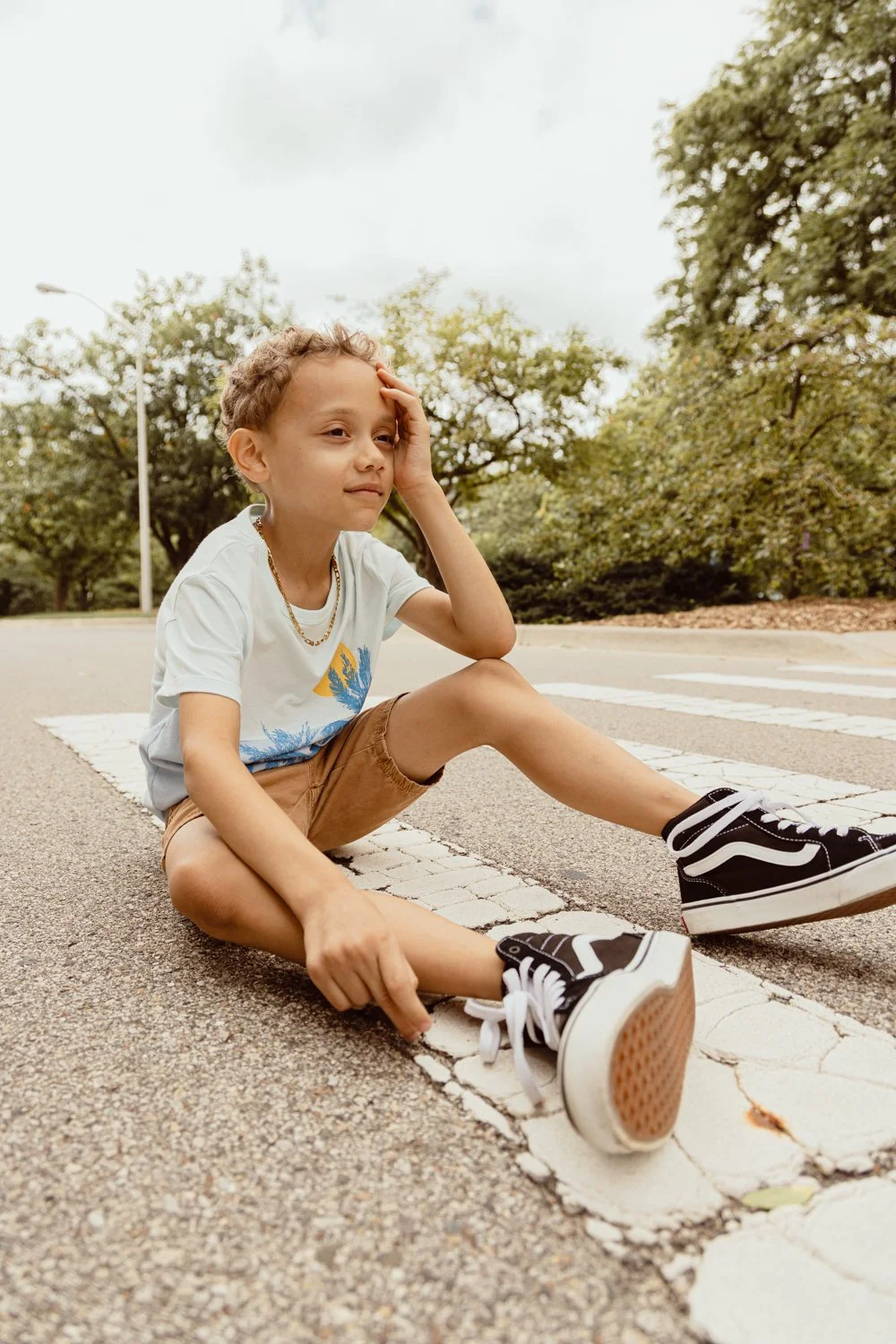 A young boy sitting on a crosswalk on the road, looking tired and holding his head, wearing a white t-shirt, tan shorts, and black sneakers, with trees and overcast sky in the background.