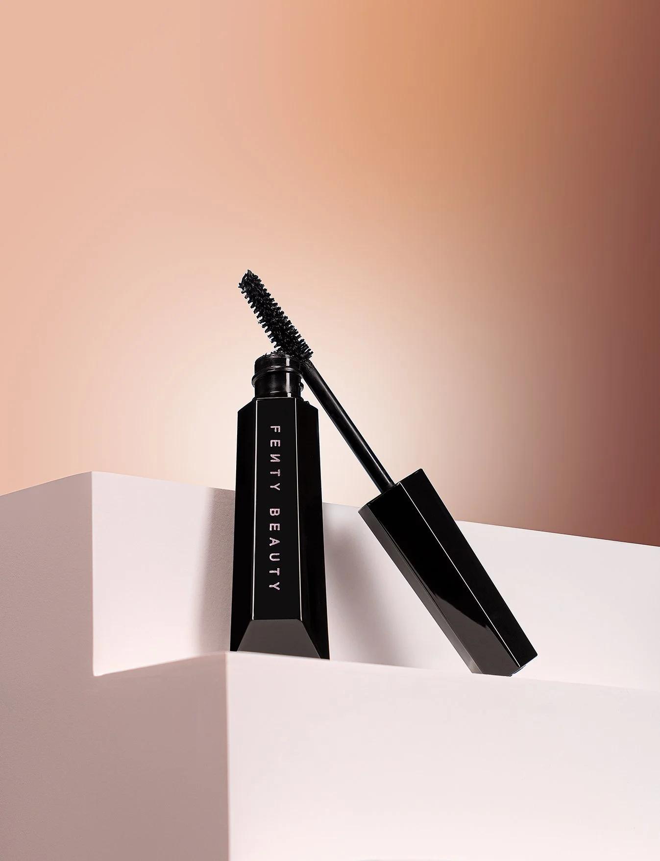 Open black mascara tube with applicator brush placed on white surface, with peach and beige background.