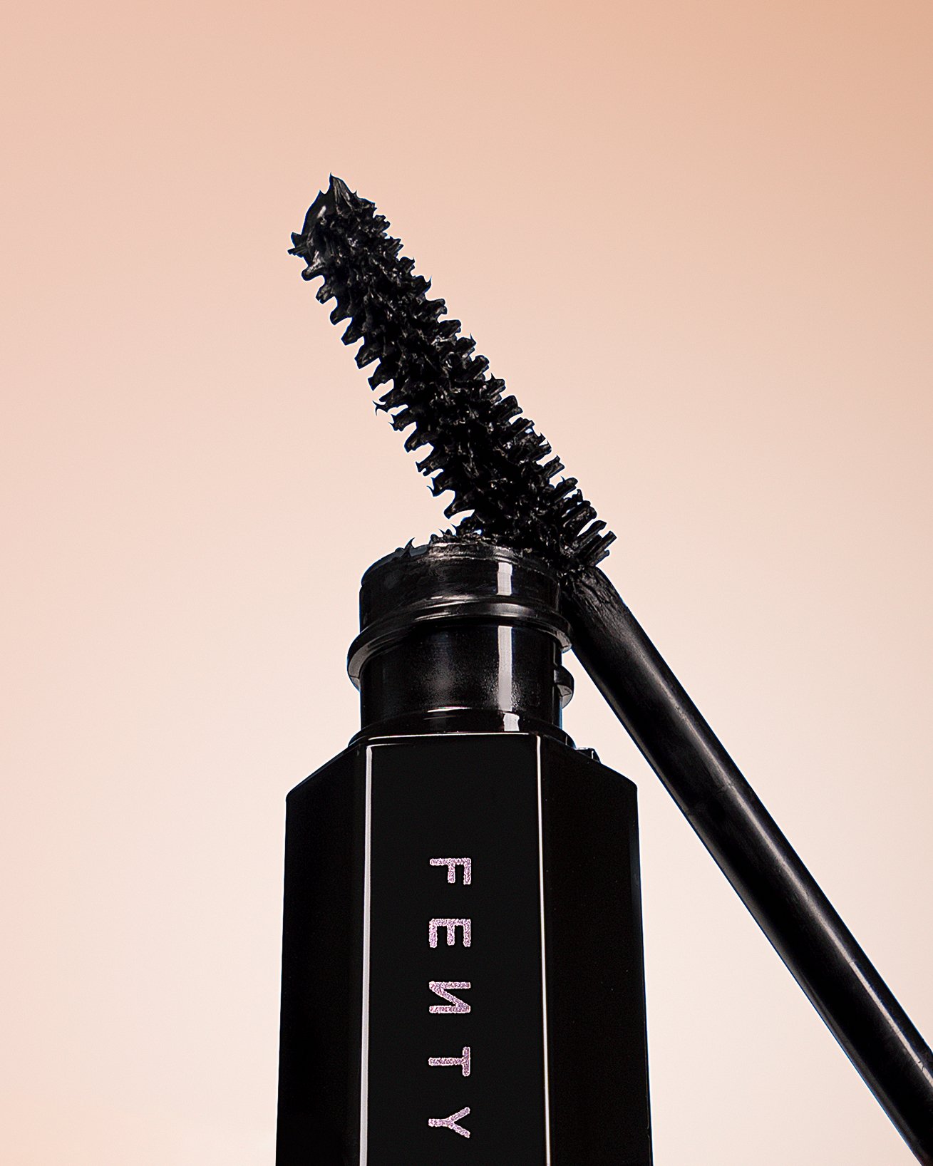 Close-up of a black mascara tube with an open mascara wand resting on top, against a plain background.