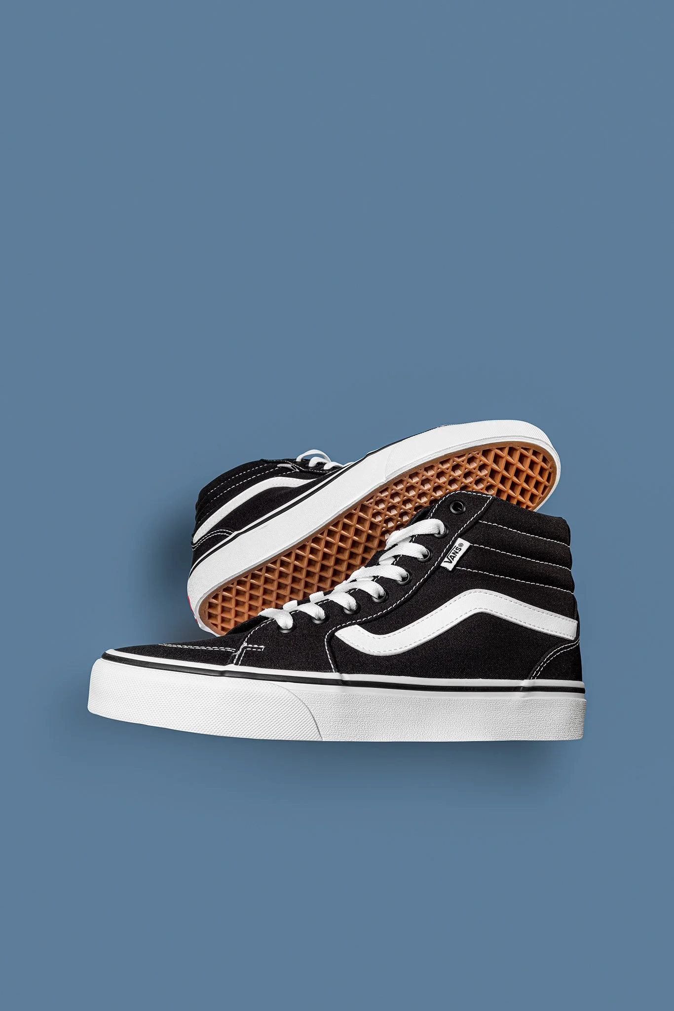 A pair of black and white Vans high-top sneakers with white laces, placed on a blue background, one shoe slightly overlapping the other, with the sole of the shoe facing up.