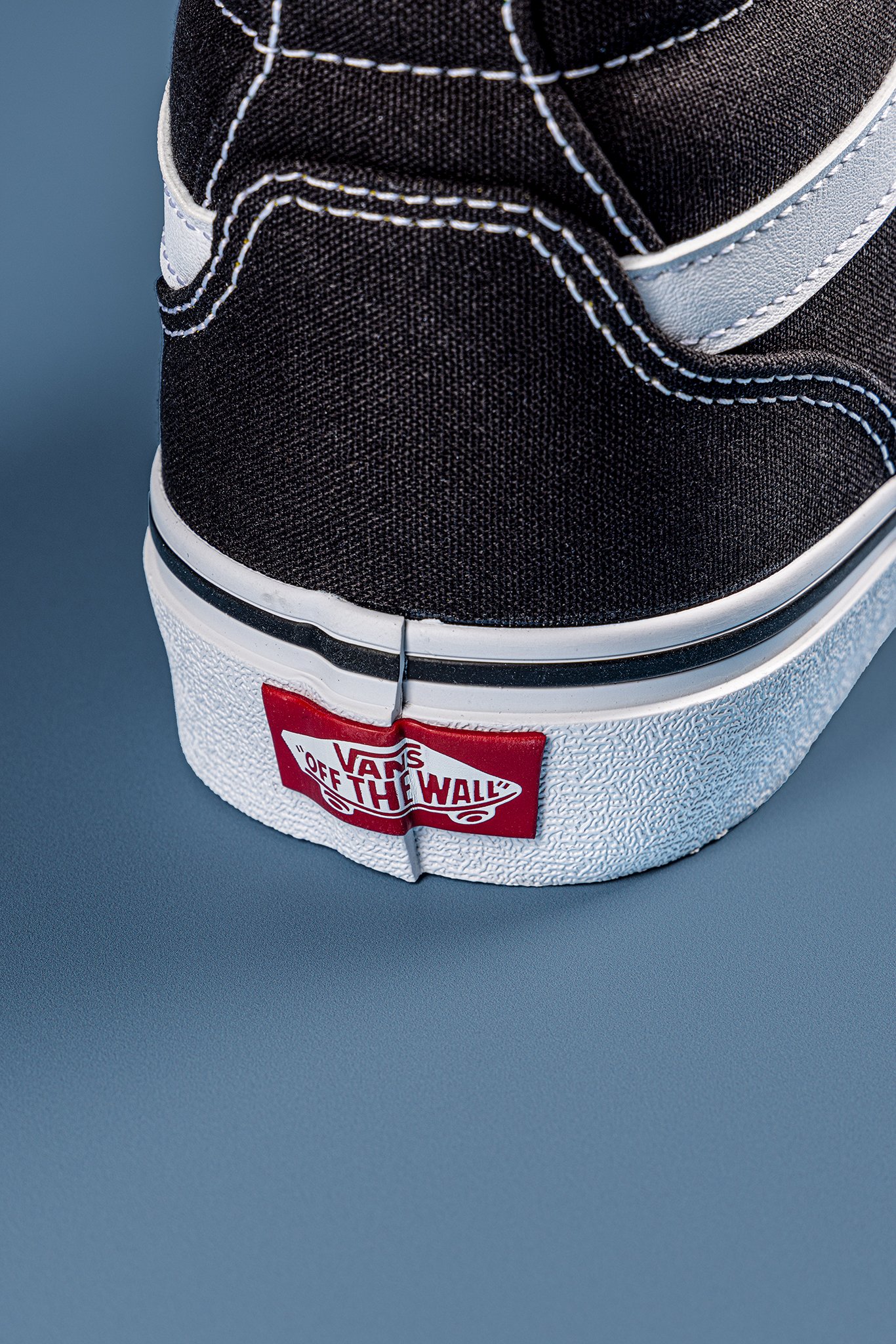 Close-up of the front part of a black Vans sneaker with white stitching and a white sole, featuring a red Vans 'Off The Wall' logo tag.