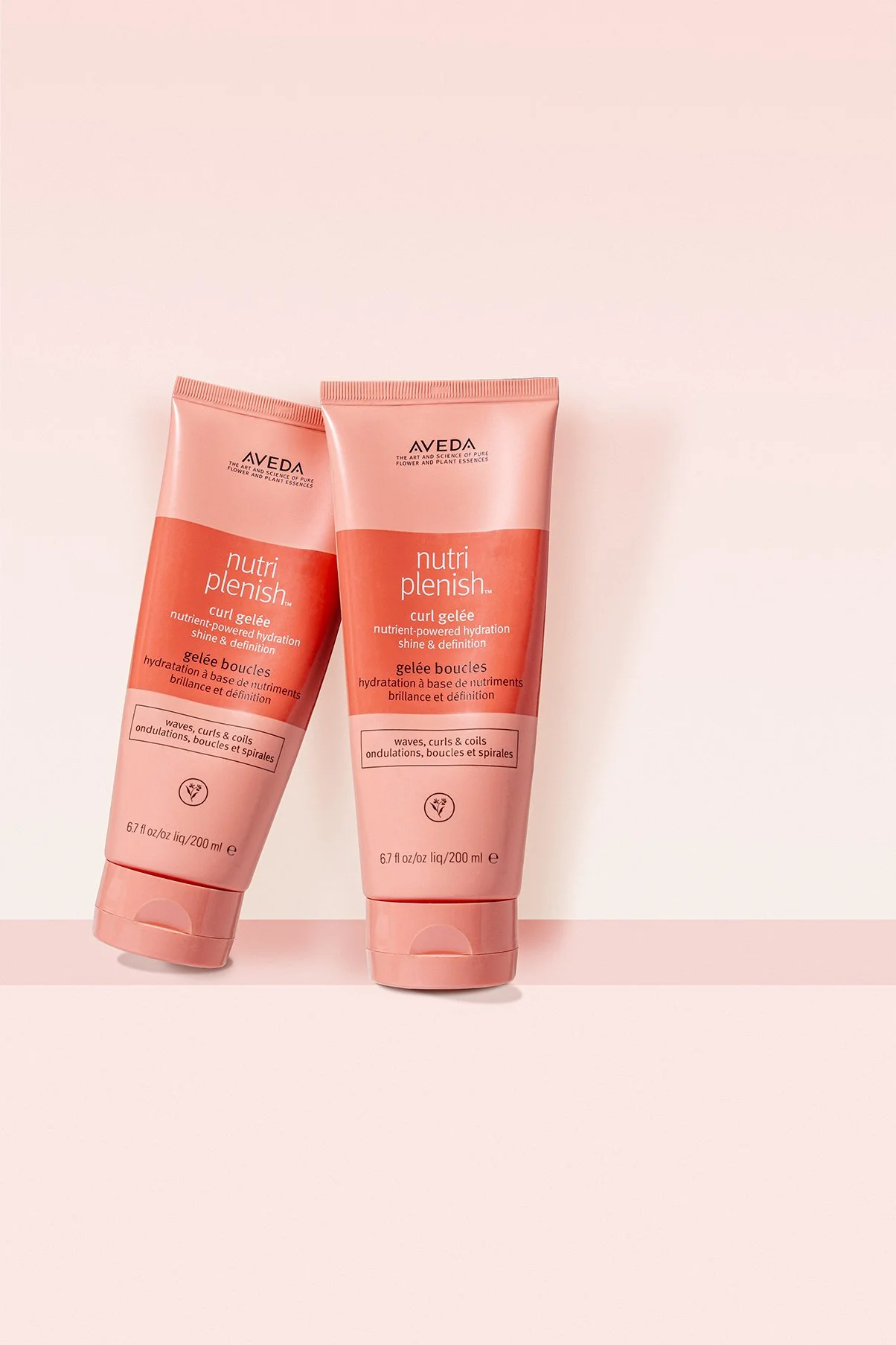 Two tubes of Aveda Nutri Plenish curl gelée standing upright on a light pink surface against a light pink background.