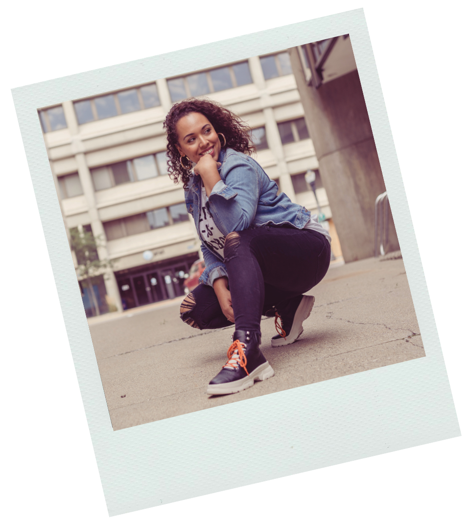 A young woman with curly hair crouching on the sidewalk outside a building, smiling and resting her chin on her hand, wearing a denim jacket, ripped black jeans, and black high-top sneakers with orange laces.