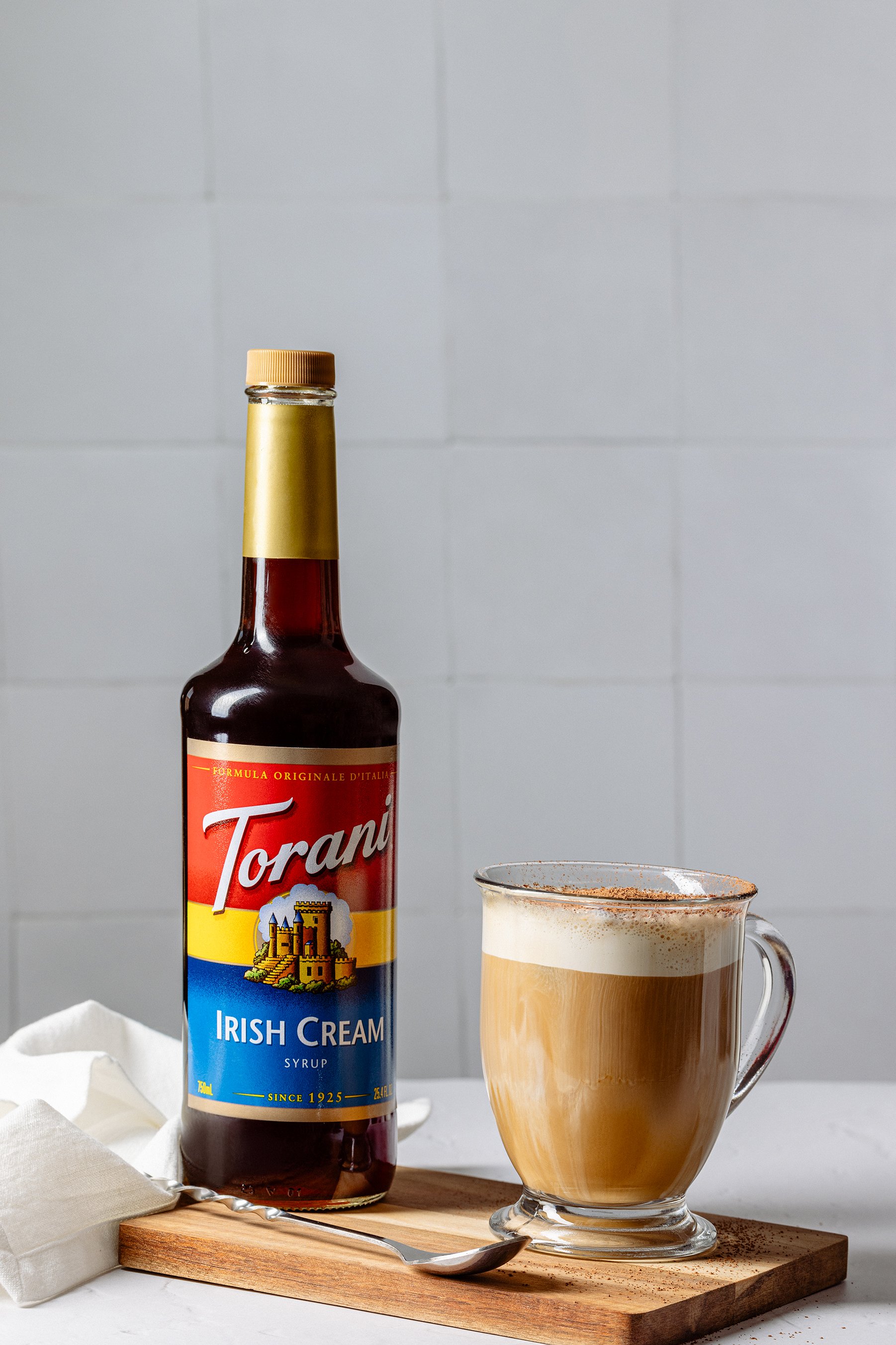 A bottle of Torani Irish Cream syrup and a glass mug of creamy coffee with foam, placed on a wooden board with a spoon and a white cloth napkin.
