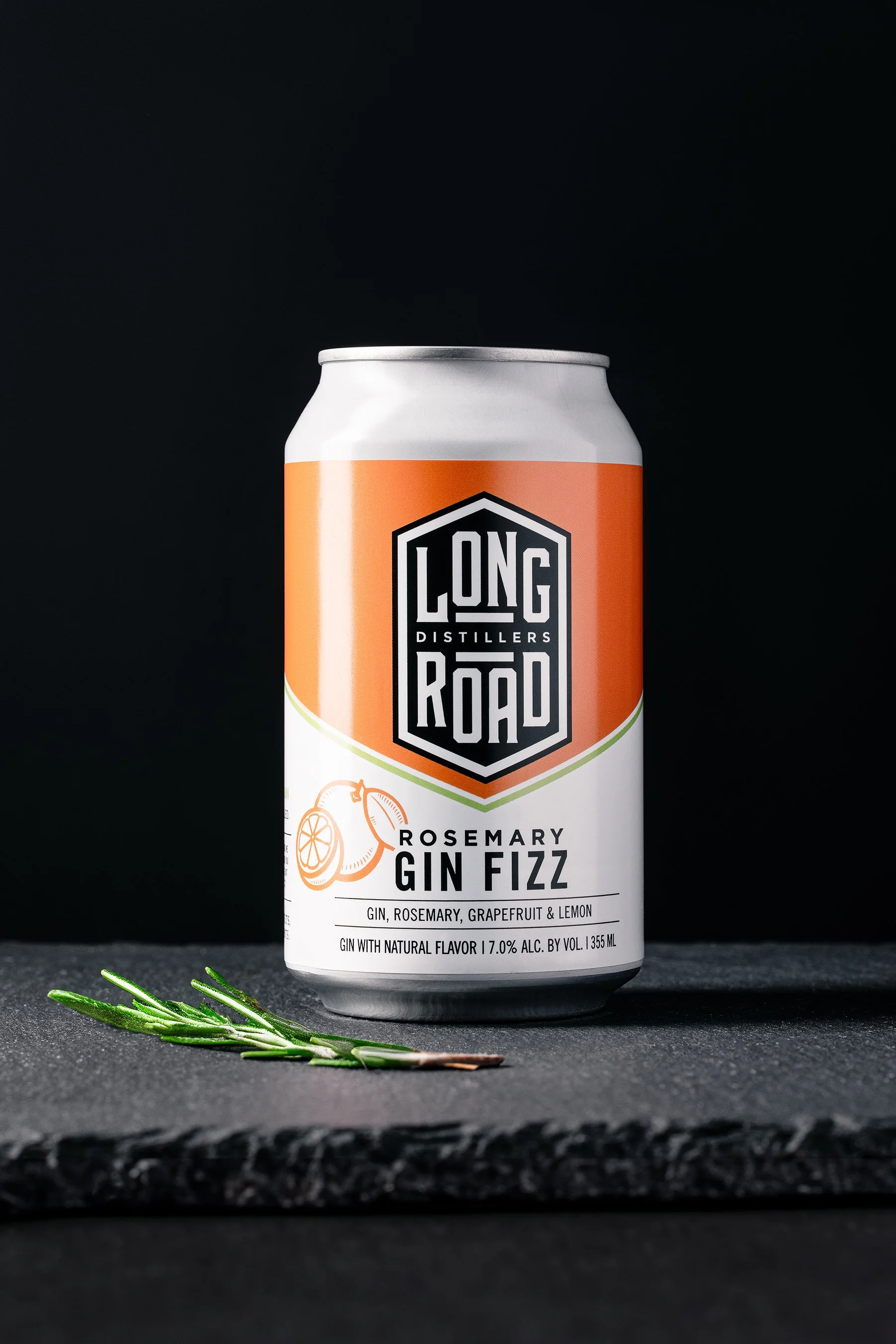 A can of Long Road Rosemary Gin Fizz with a sprig of rosemary in front of it, on a dark surface with a dark background.