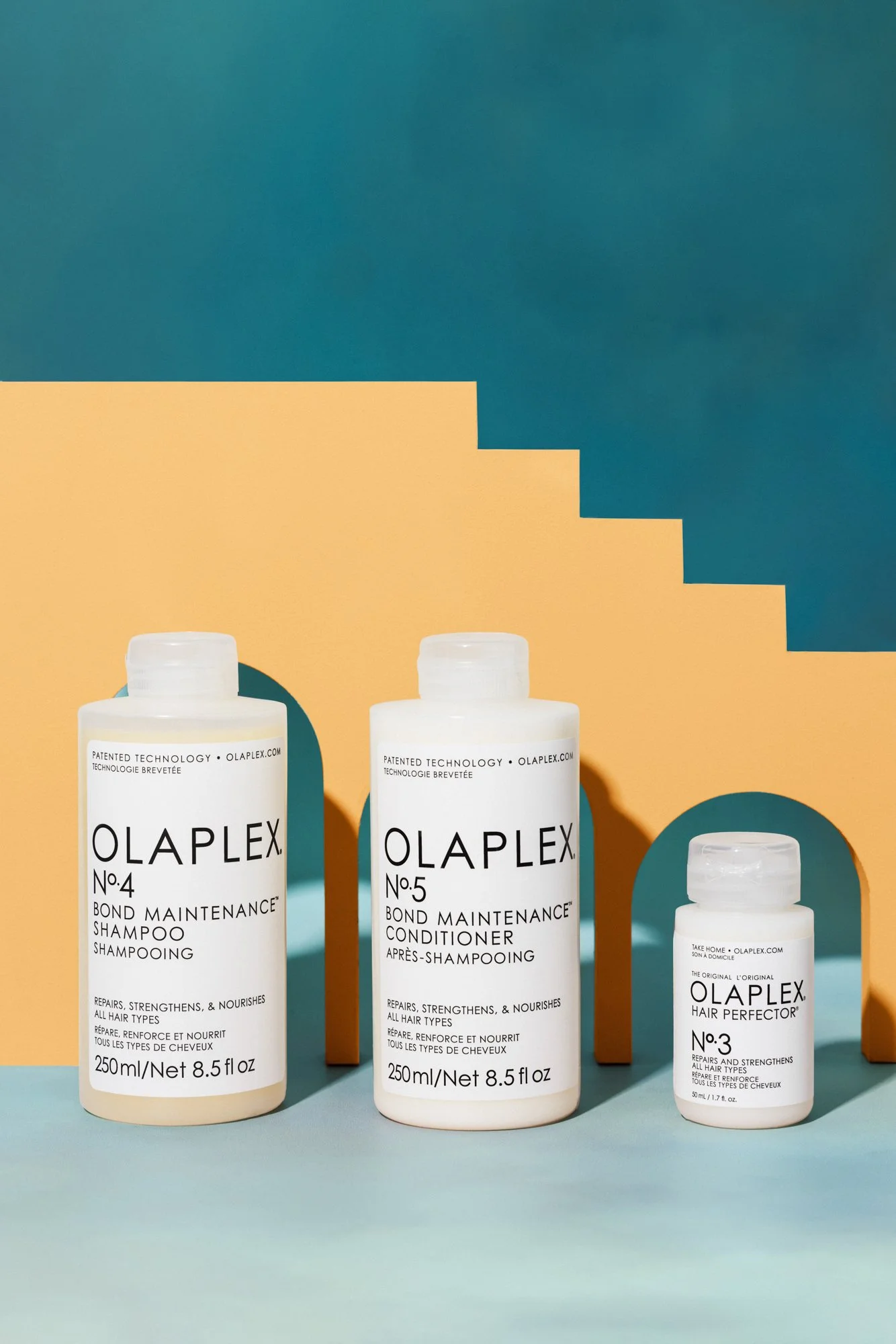 Three bottles of Olaplex hair care products, including No. 4 Bond Maintenance Shampoo, No. 5 Bond Maintenance Conditioner, and No. 3 Hair Perfector, arranged on a colorful background with blue, yellow, and teal tones.