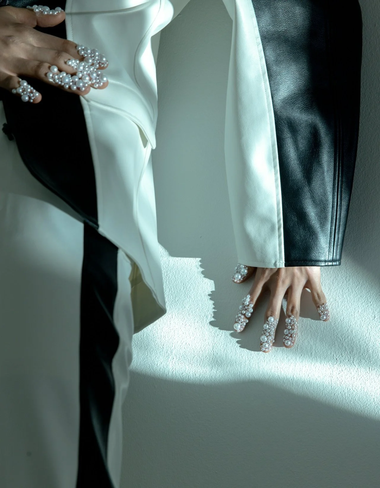 Close-up of a person's hand resting on a wall, wearing a pearl ring and pearl-decorated nails. The person is dressed in a white jacket with black and white leather pants, illuminated by natural light.