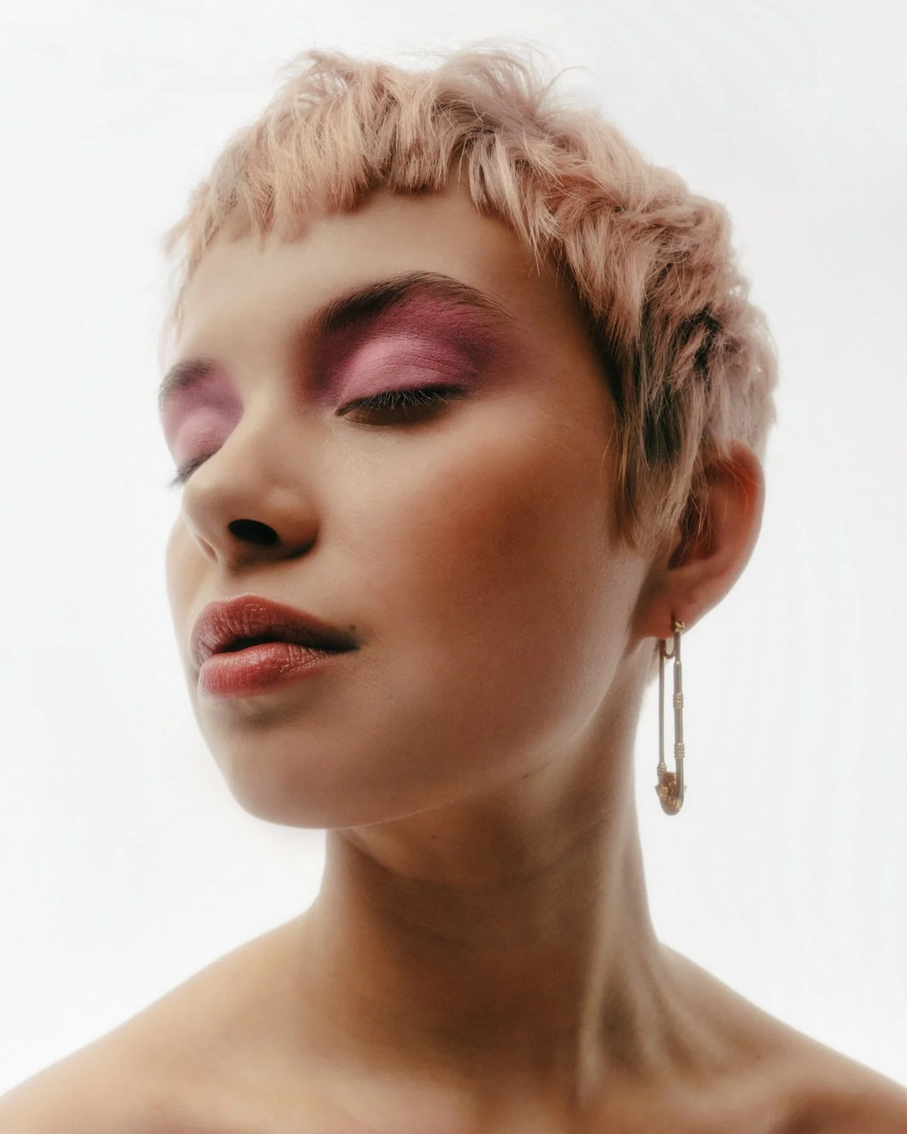 Close-up of a woman with short pink hair, pink eyeshadow, and wearing an asymmetric earring, with her eyes closed.