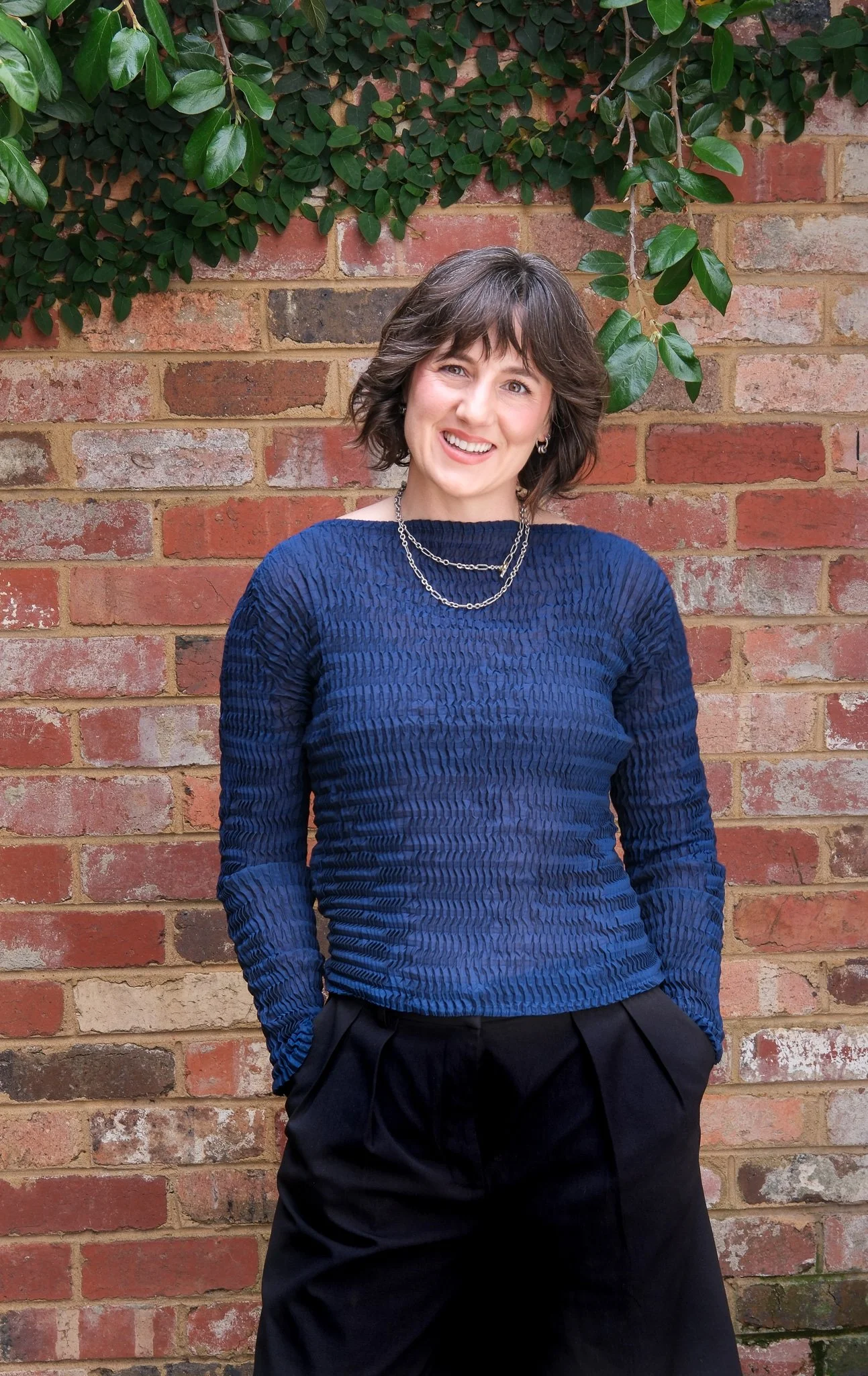 A woman with short brown hair, wearing a textured blue long-sleeve top and black pants, smiling while standing outdoors in front of a brick wall with green foliage overhead.
