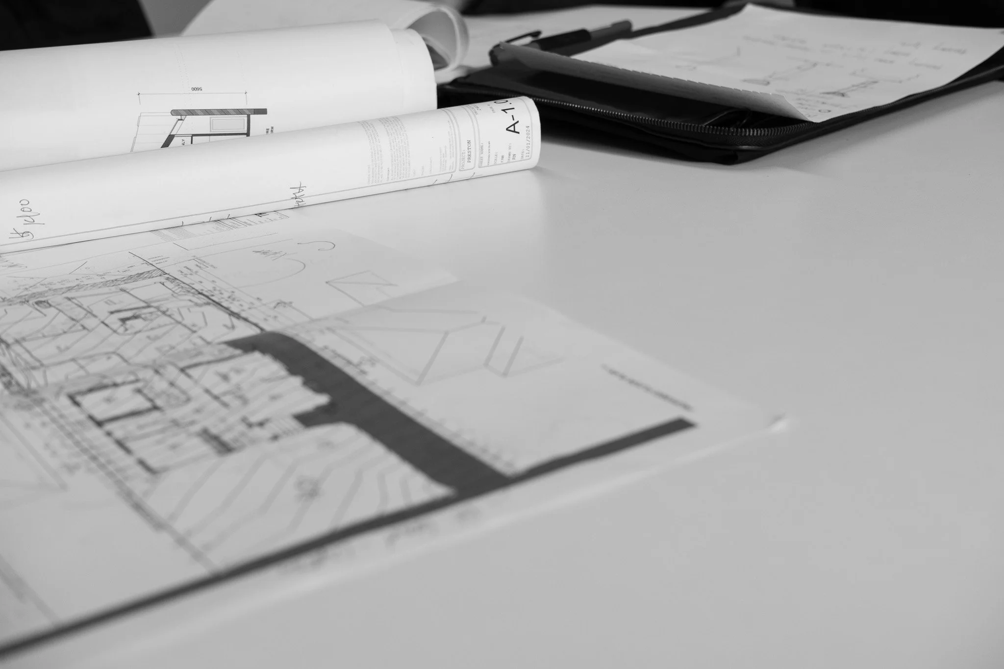 Architectural blueprints and documents spread out on a desk, with some rolled-up plans and a folder in the background.