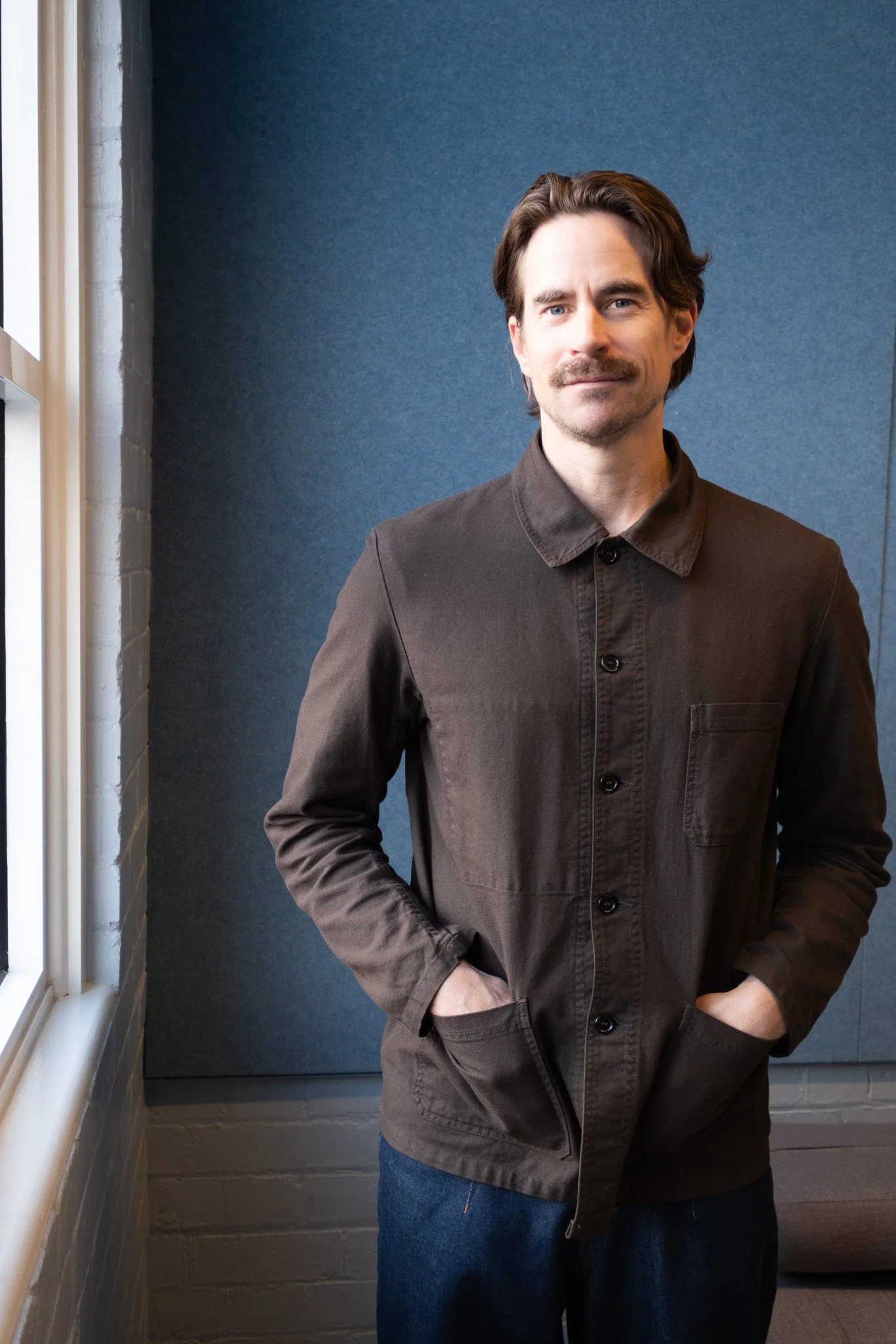 A man with dark hair and a mustache wearing a dark brown button-up jacket, standing with hands in pockets near a window and a blue wall background.