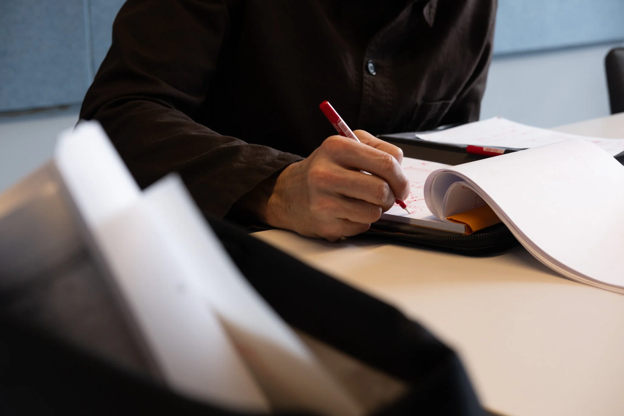 A person writing with a red pen on a sheet of paper in a spiral notebook, placed on a desk with additional papers and a backpack visible in the foreground.