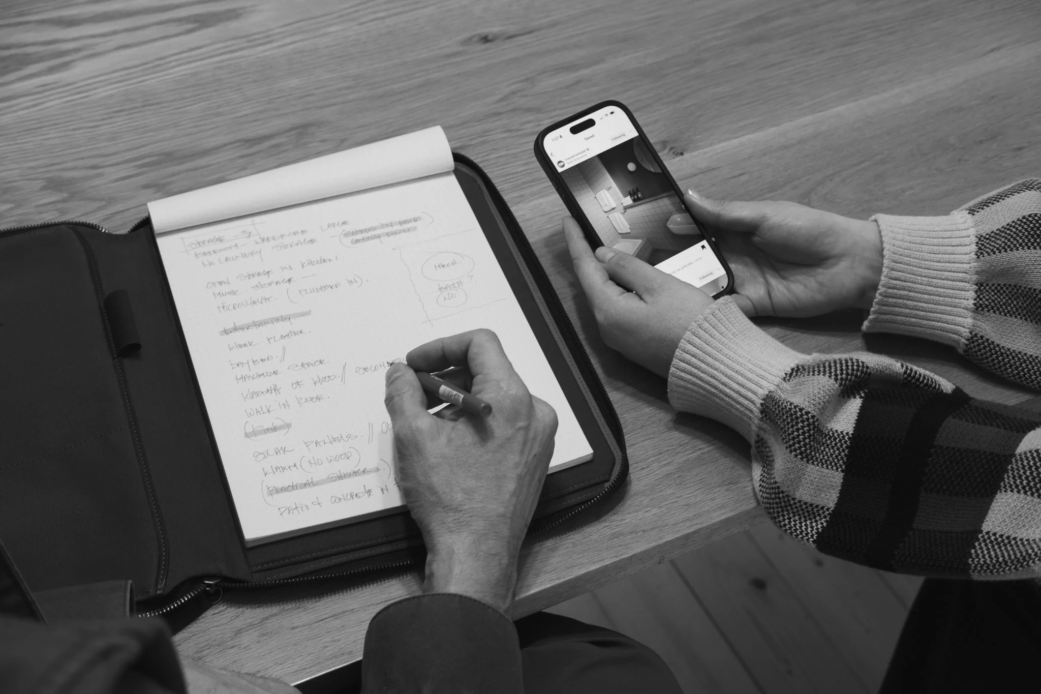 Person holding a smartphone and writing notes in a notebook at a wooden desk.