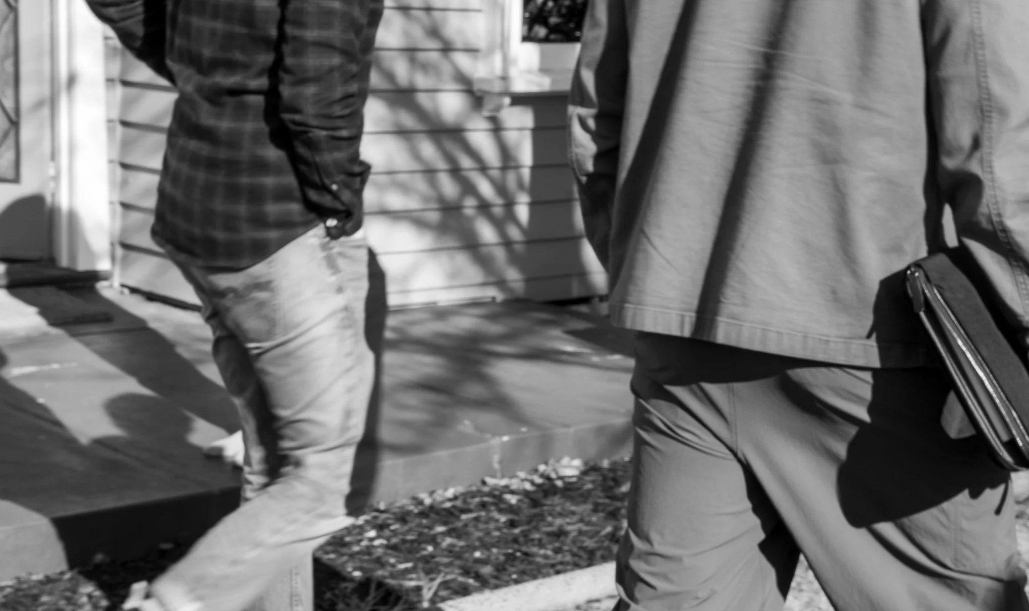 Two men walking outdoors, one in a checkered shirt and the other in a plain shirt with a bag, on a sidewalk with some shadows.