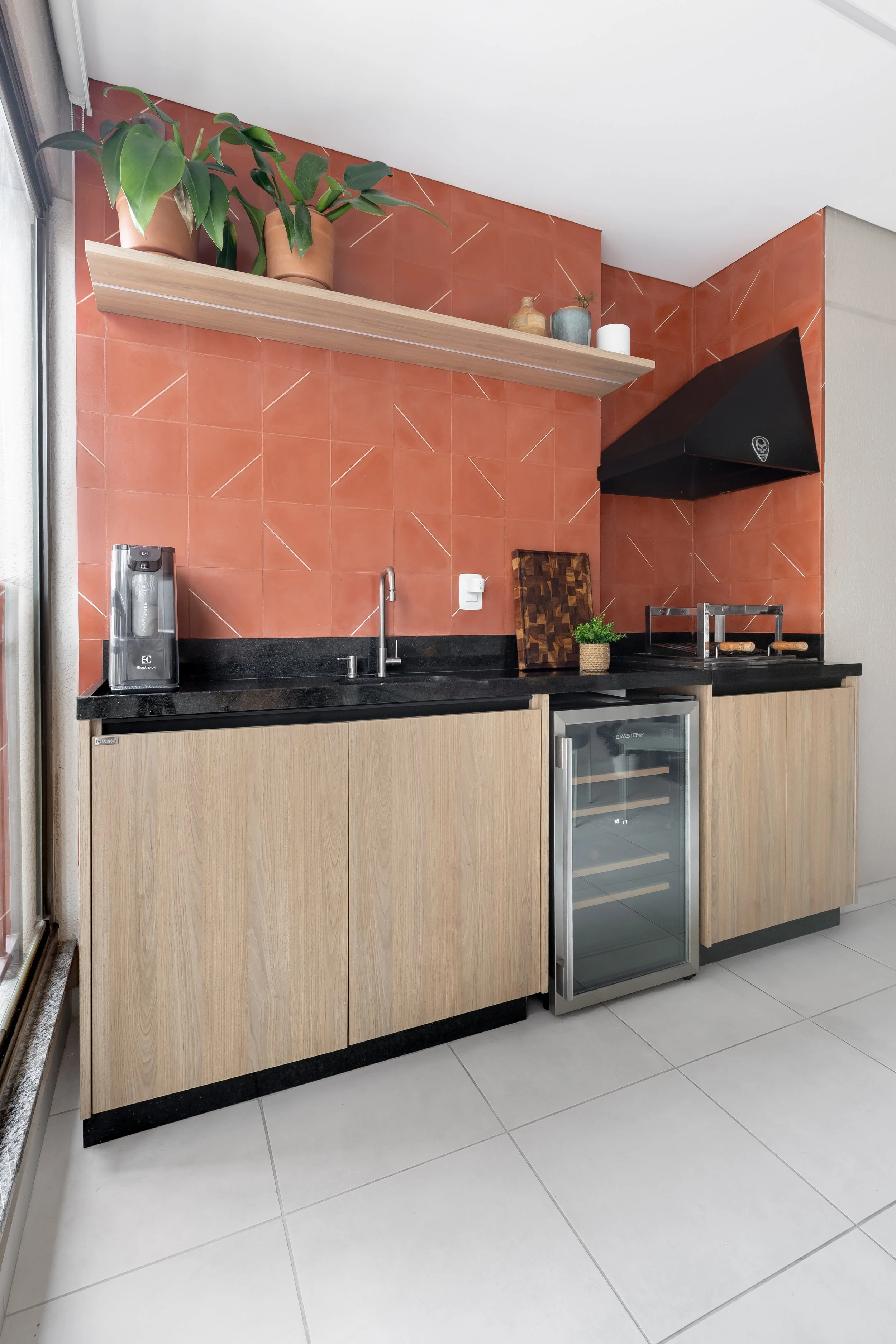 Cozinha moderna com armários de madeira clara, parede de azulejos laranja, bancada preta, pia, três plantas em vasos na prateleira, e um exaustor preto acima do fogão.