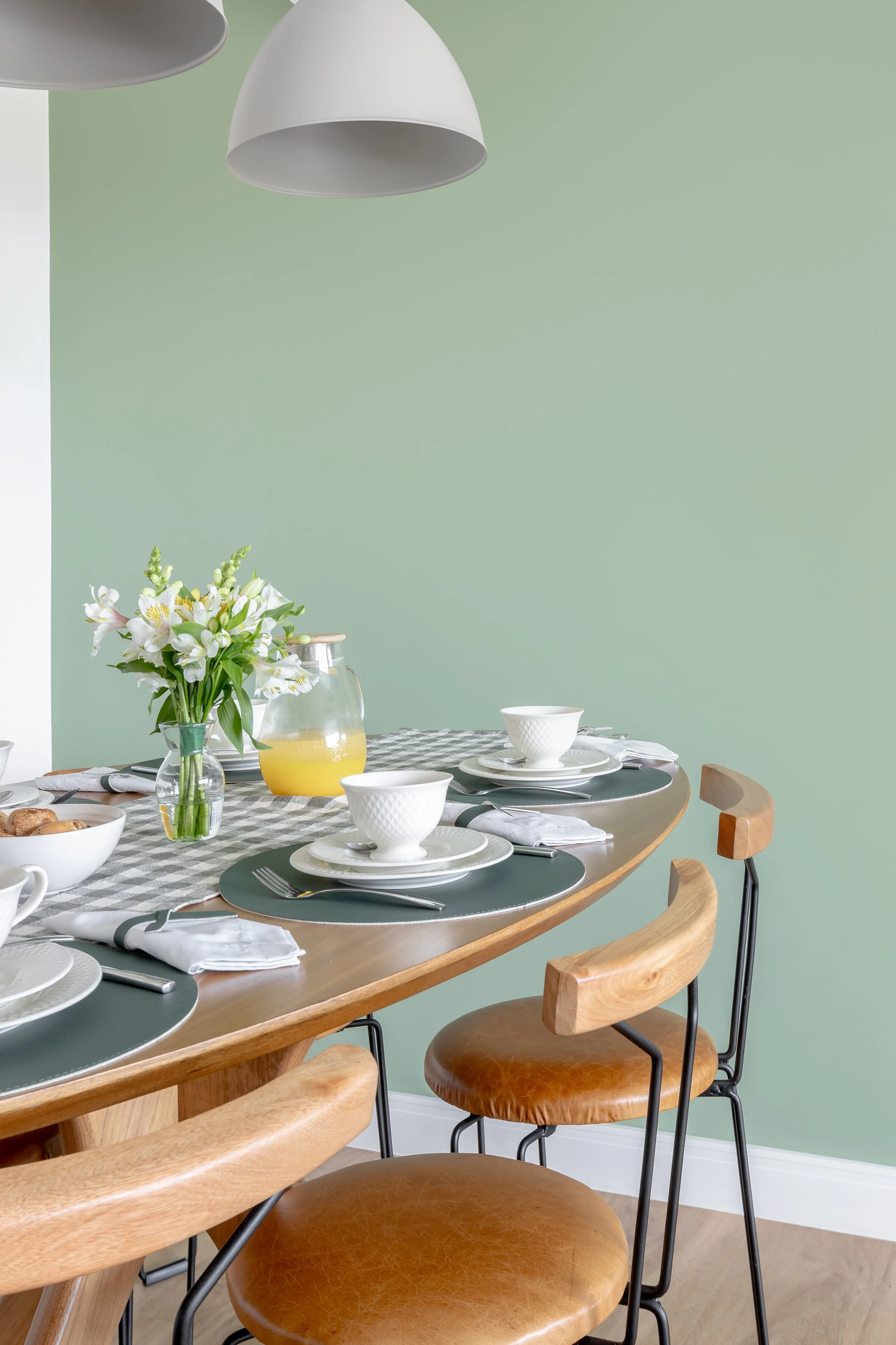 Mesa de jantar com louças brancas, guardanapos brancos, talheres, uma jarra de suco, um vaso com flores e cadeiras de madeira com assentos de couro. Fundo verde claro.