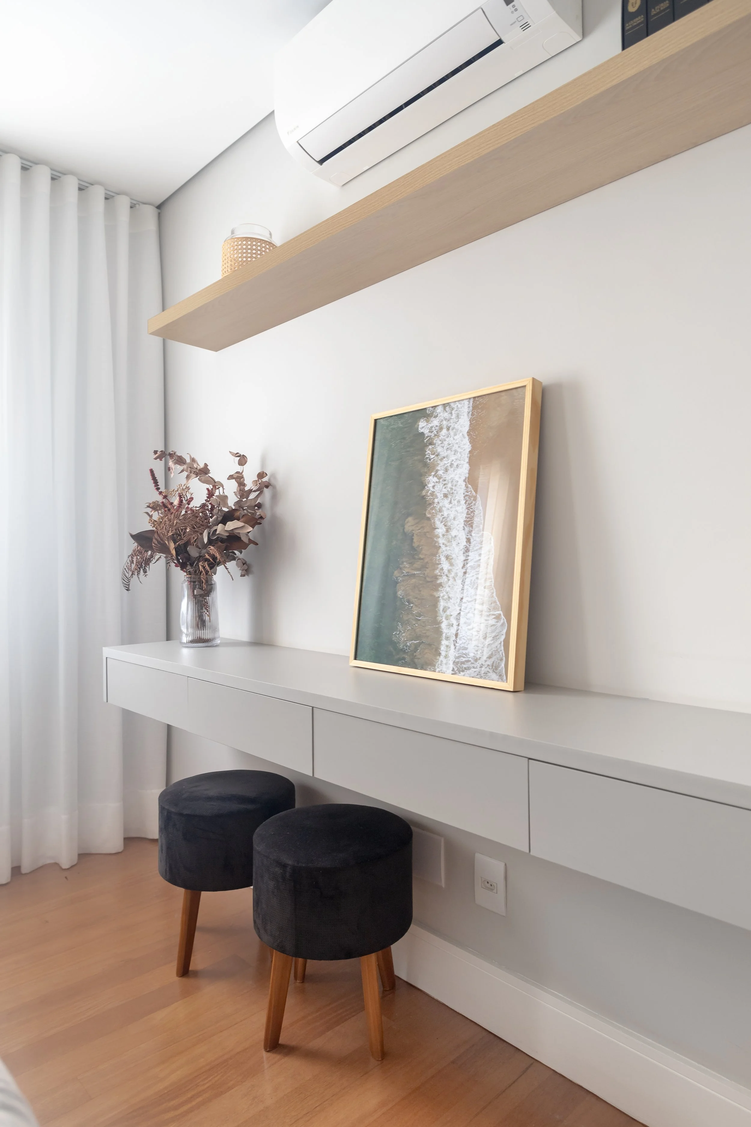 Sala com mesa branca, duas banquetas pretas com pernas de madeira, uma planta seca em vaso de vidro, quadro de paisagem de praia encostado na parede, prateleira de madeira com objeto decorativo, ar-condicionado na parede, cortinas brancas ao lado da 