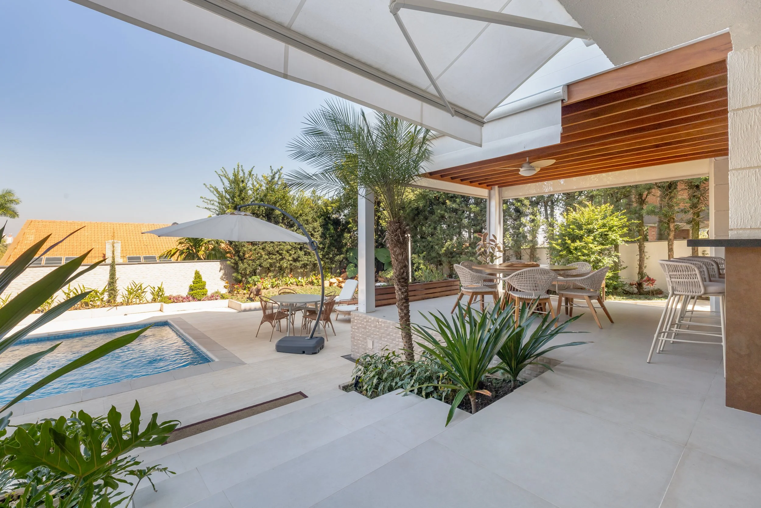 Varanda com área de churrasqueira, mesa com cadeiras, piscina pequena, plantas e árvores, sombra de guarda-sol e céu claro.