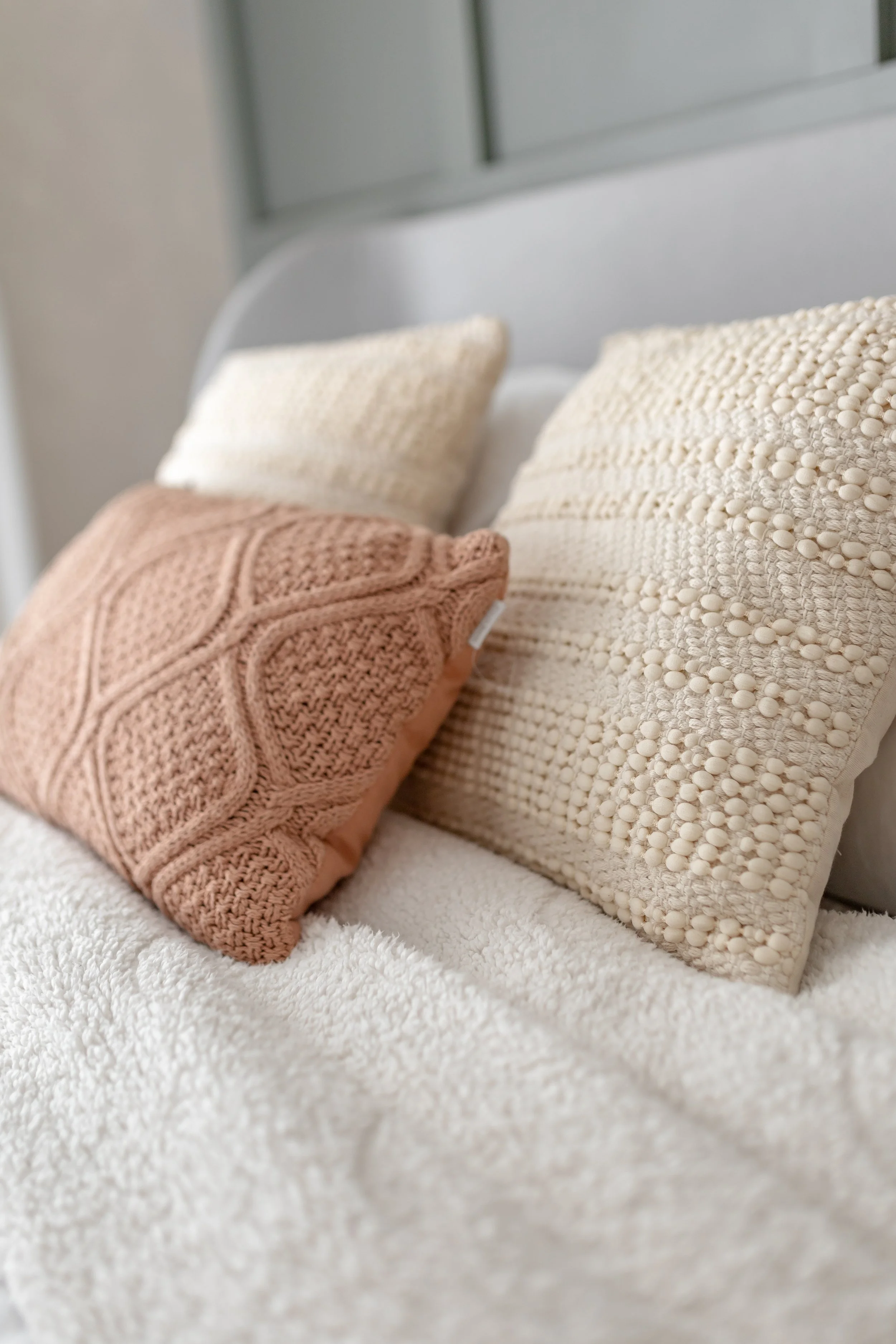 Três almofadas de tricô, sendo uma cor de rosa e duas de creme, colocadas sobre uma cama com lençol branco. Fundo desfocado de parede cinza.