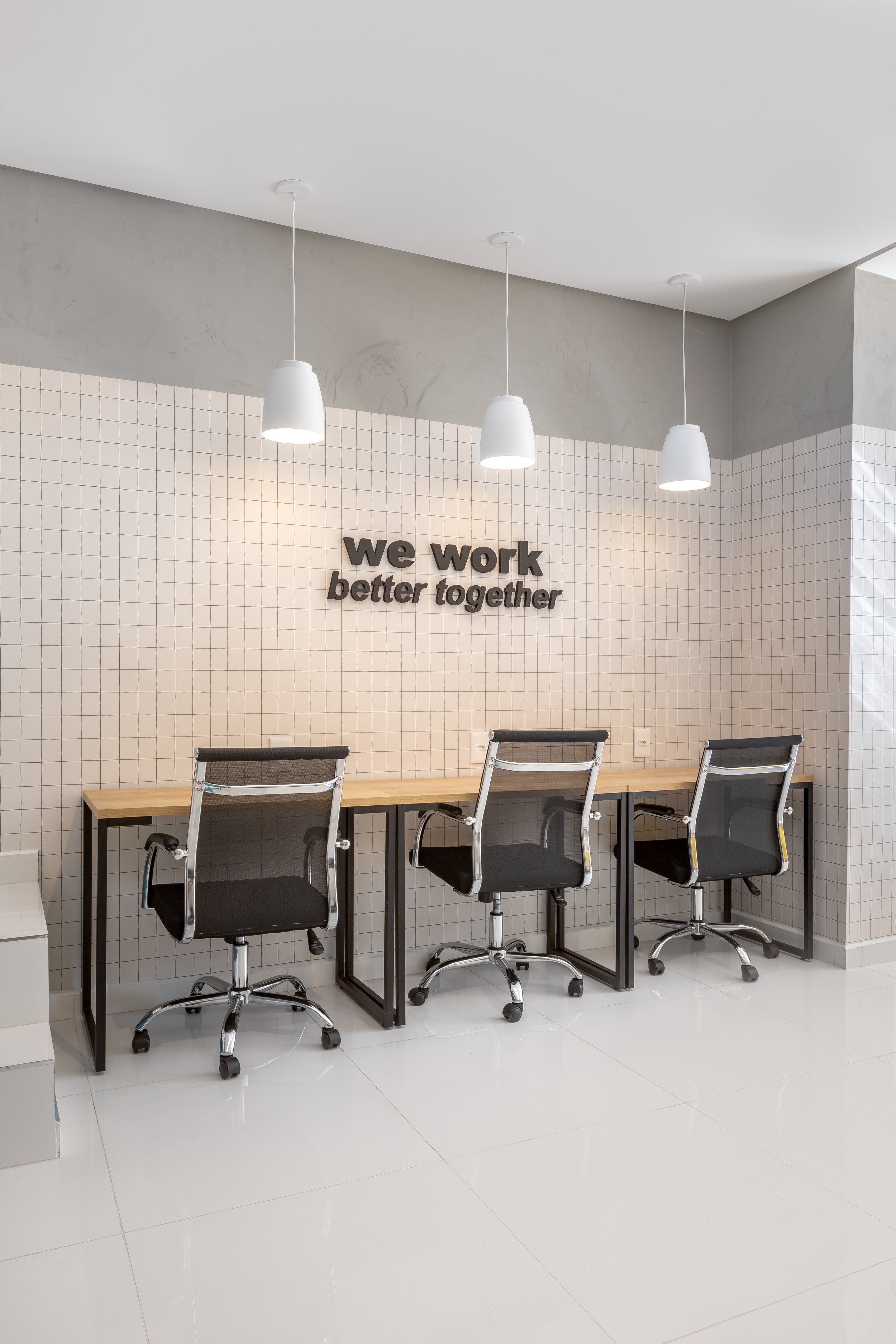 Três cadeiras de escritório pretas com estrutura metálica e rodízios, frente para uma mesa de madeira, em um ambiente de escritório moderno com paredes de azulejos brancos e uma placa com a frase "we work better together" ao fundo.