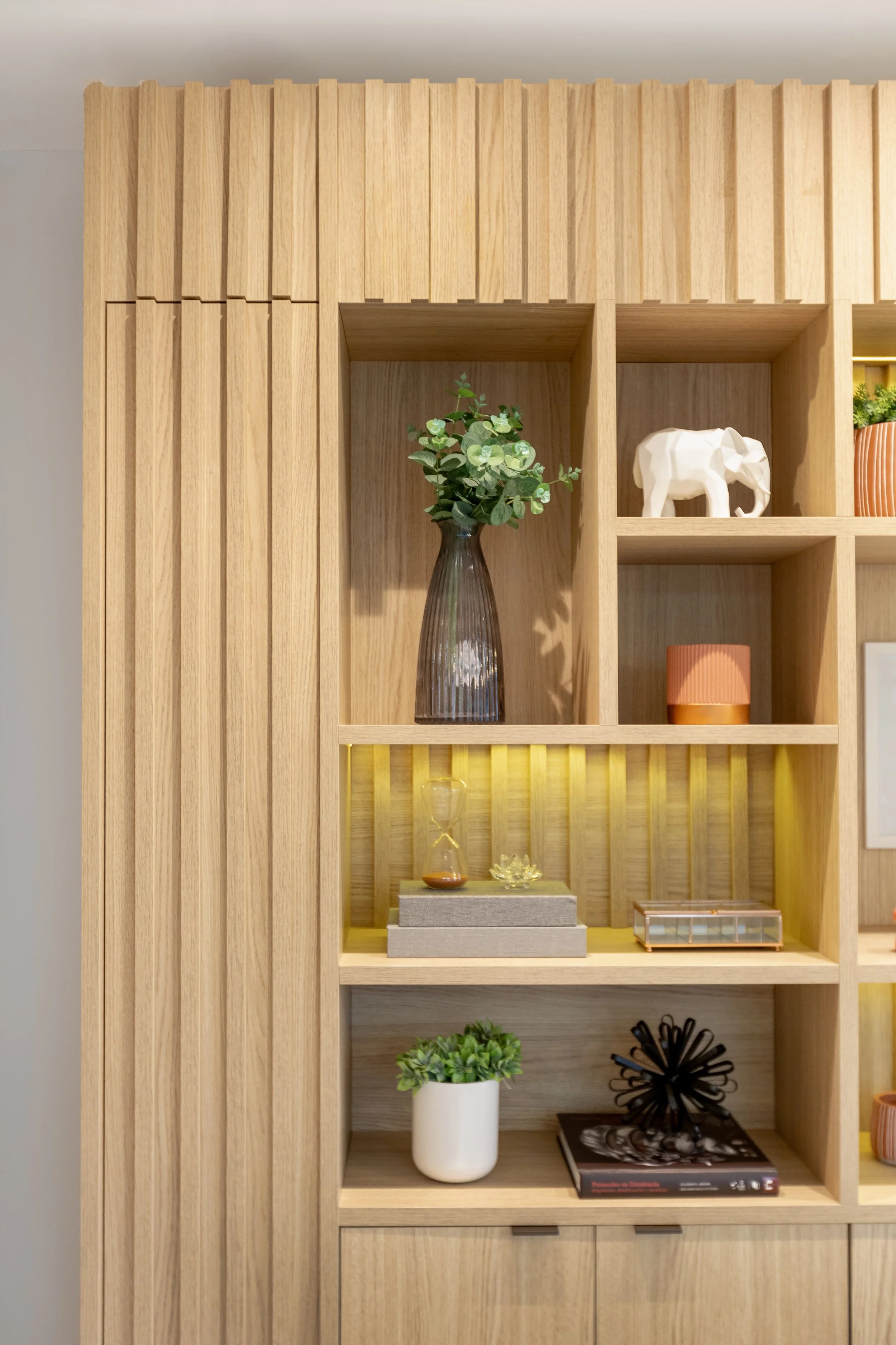 Estante de madeira com várias prateleiras decorativas, contendo plantas, uma estatueta de elefante branca, livros, uma caixa decorativa, uma luminária laranja, e um vaso de planta com folhagem verde.