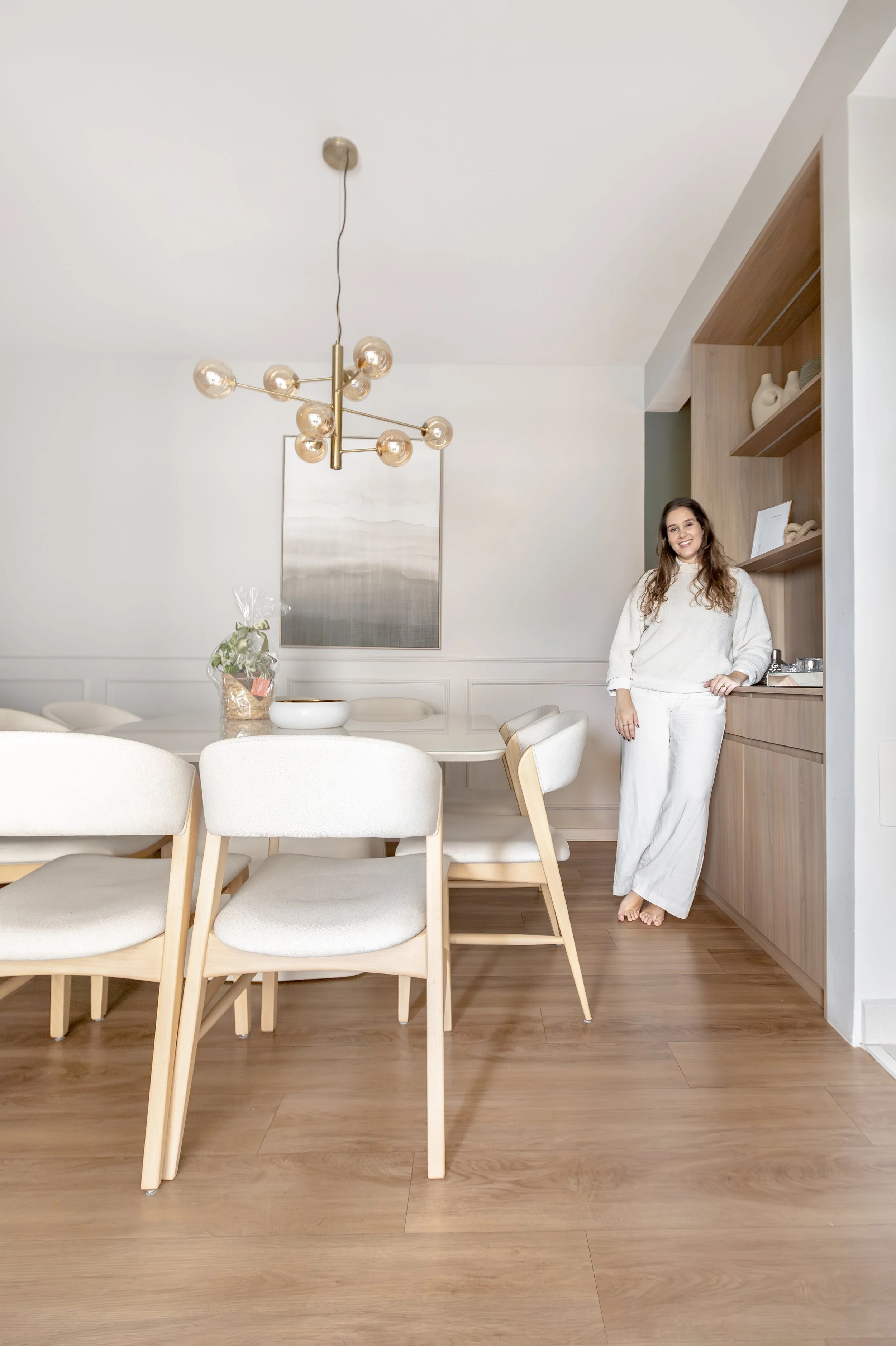 Mulher sorridente com cabelo castanho claro, usando suéter branco e calça branca, posando ao lado de uma parede de madeira. A sala tem uma mesa de jantar branca, cadeiras brancas com estrutura de madeira, uma luminária moderna com vários bulbos de vi