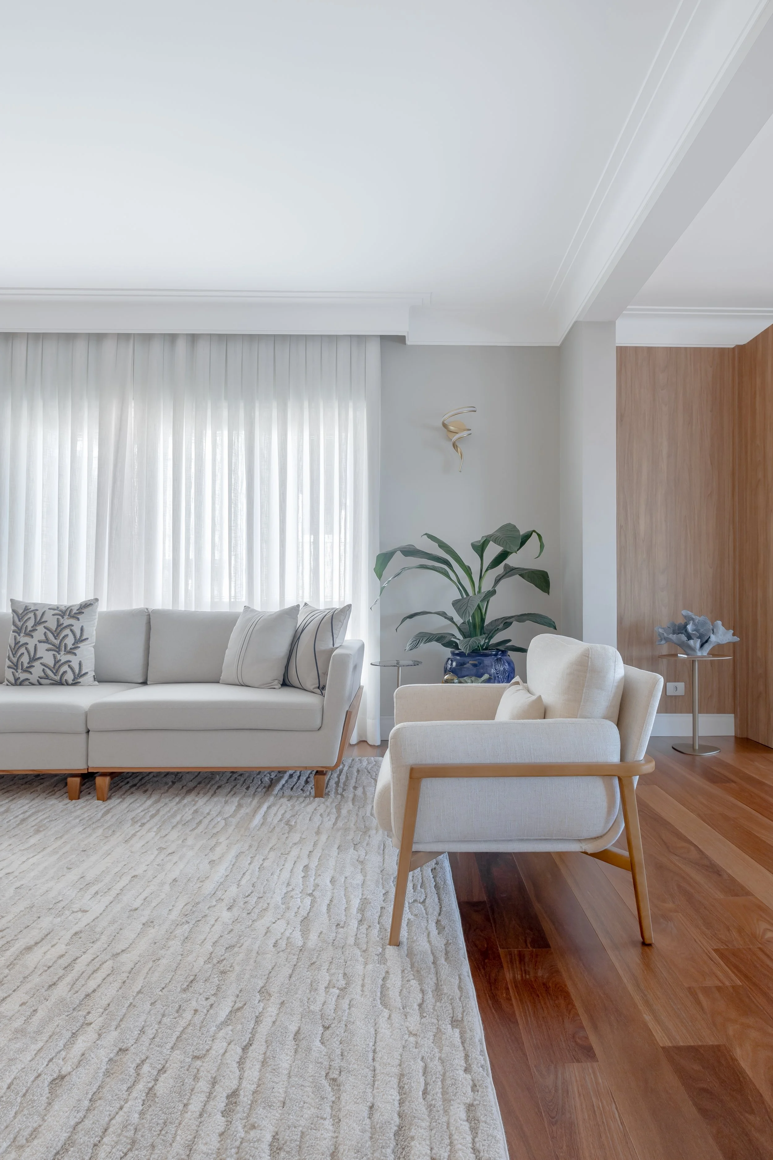 Sala de estar moderna com sofá branco, poltronas de tecido claro, cortinas brancas, tapete bege, planta grande em vaso azul, paredes brancas e piso de madeira.