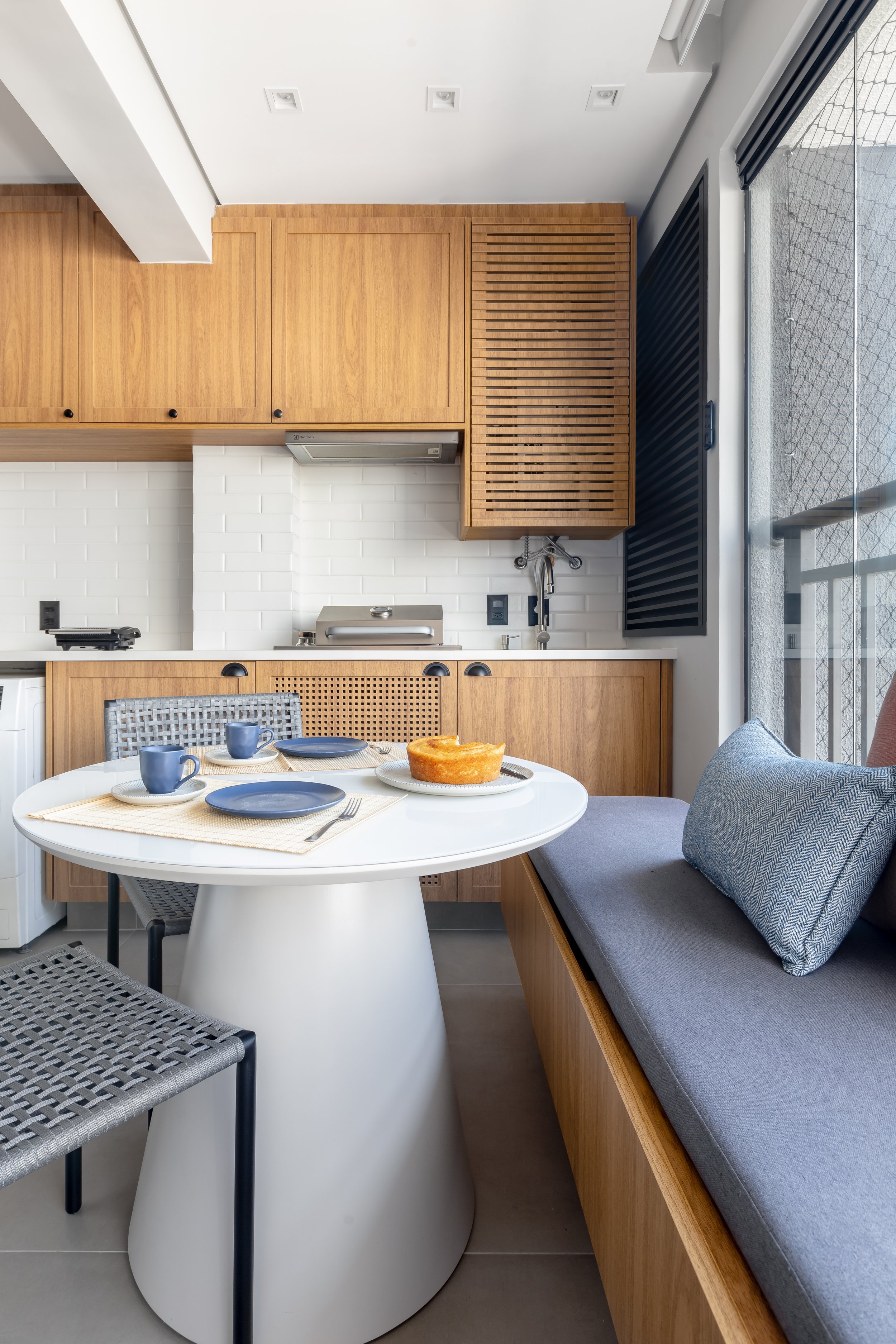 Cozinha moderna com armários de madeira, balcão branco, mesa redonda branca com louças azuis e bolo, cadeira de palha, banco de encosto com almofada azul, parede de tijolos brancos e grande janela de vidro permitindo entrada de luz natural.