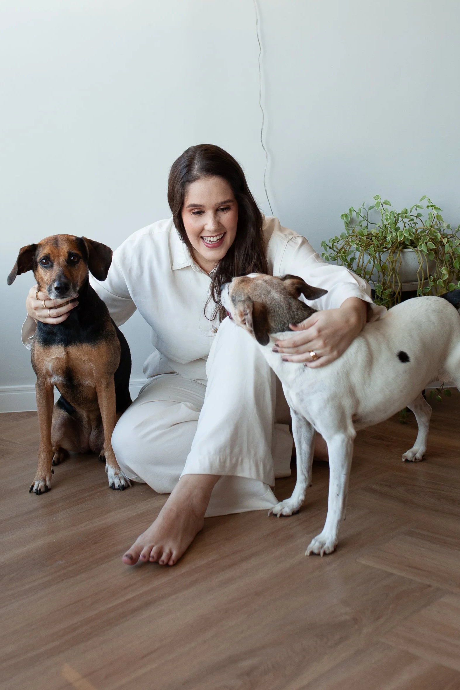 Mulher sorridente brincando com duas cachorras dentro de casa, com uma planta ao fundo e piso de madeira.