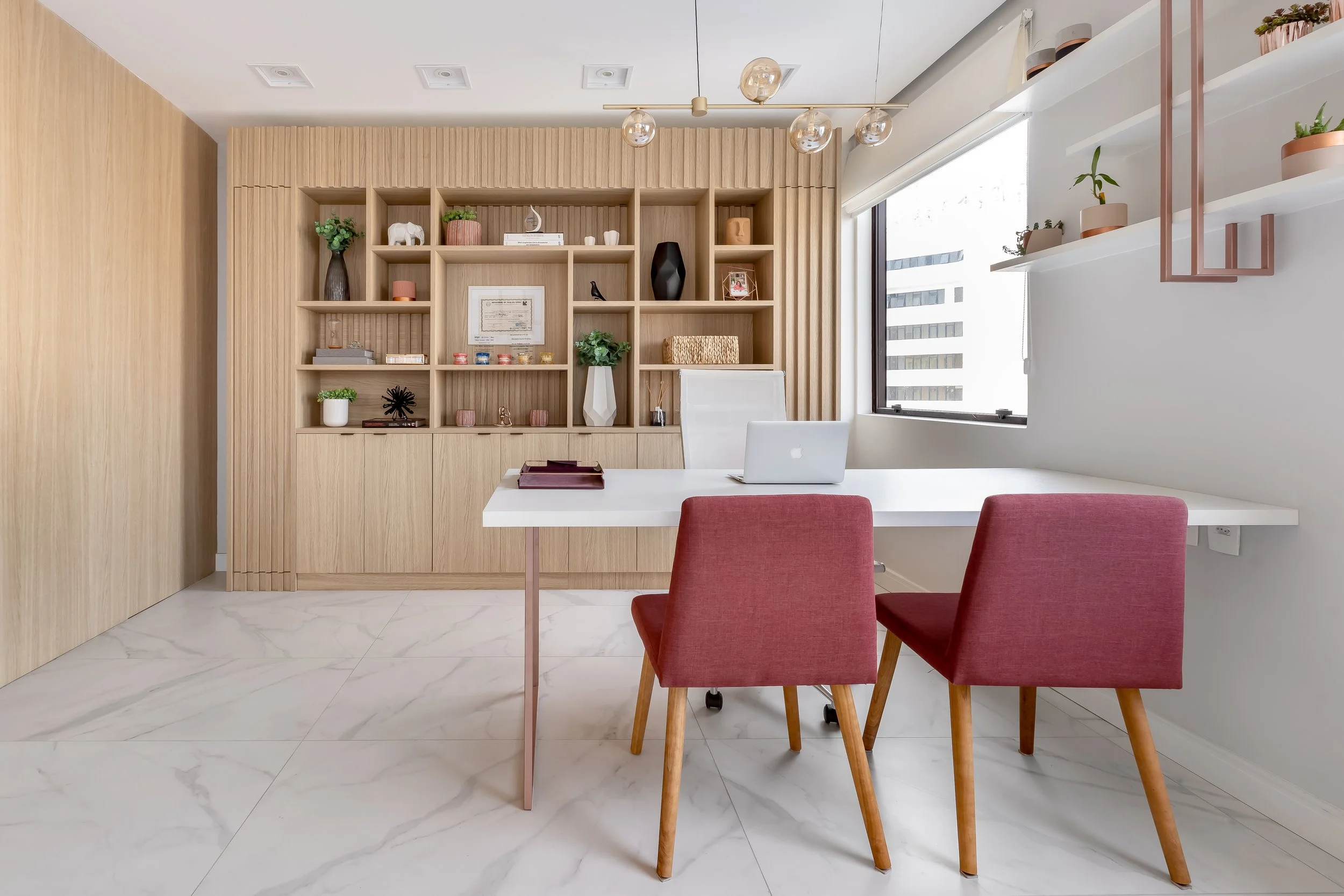 Escritório com mesa branca, duas cadeiras rosa, prateleira de madeira com objetos decorativos e uma janela grande