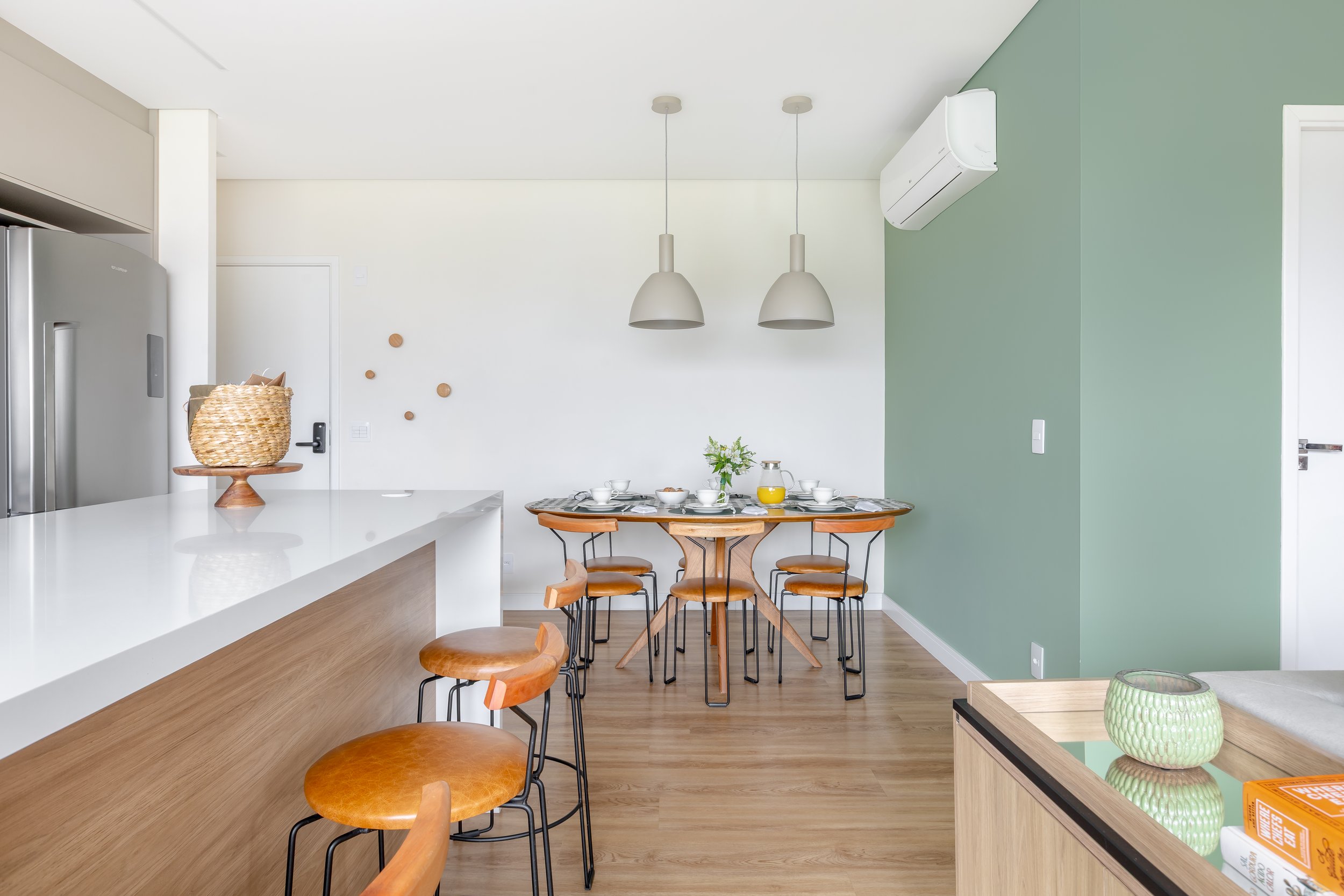 Sala de jantar moderna com mesa de madeira, cadeiras de ferro, utensílios, e duas luminárias pendentes. Cozinha com ilha de mármore e cadeiras, parede verde, ar-condicionado, e decoração minimalista.