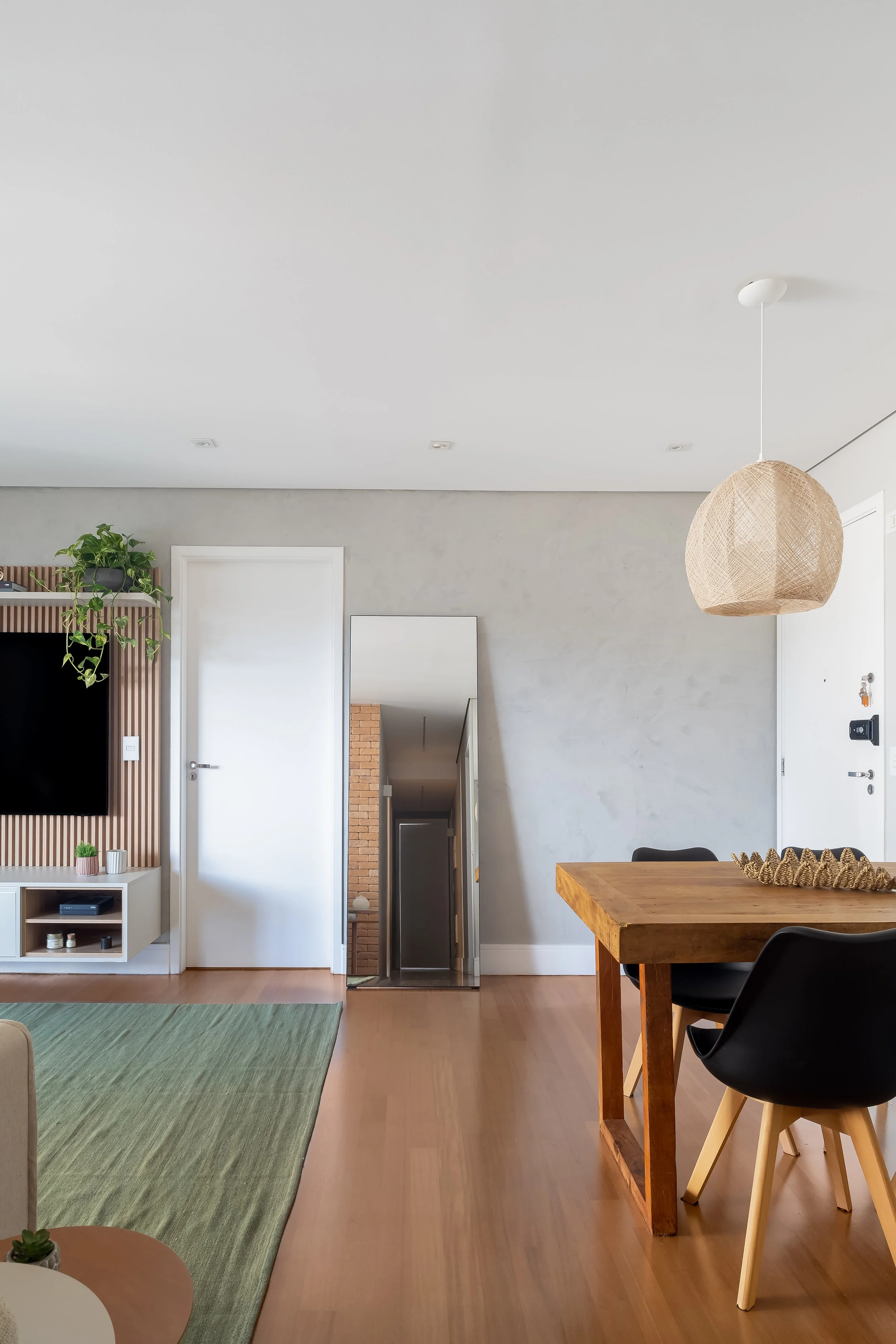 Sala de estar com mesa de jantar de madeira, cadeiras pretas e cadeira de encosto em branco, parede com espelho grande, TV na parede com painel de madeira, parede clara e piso de madeira.