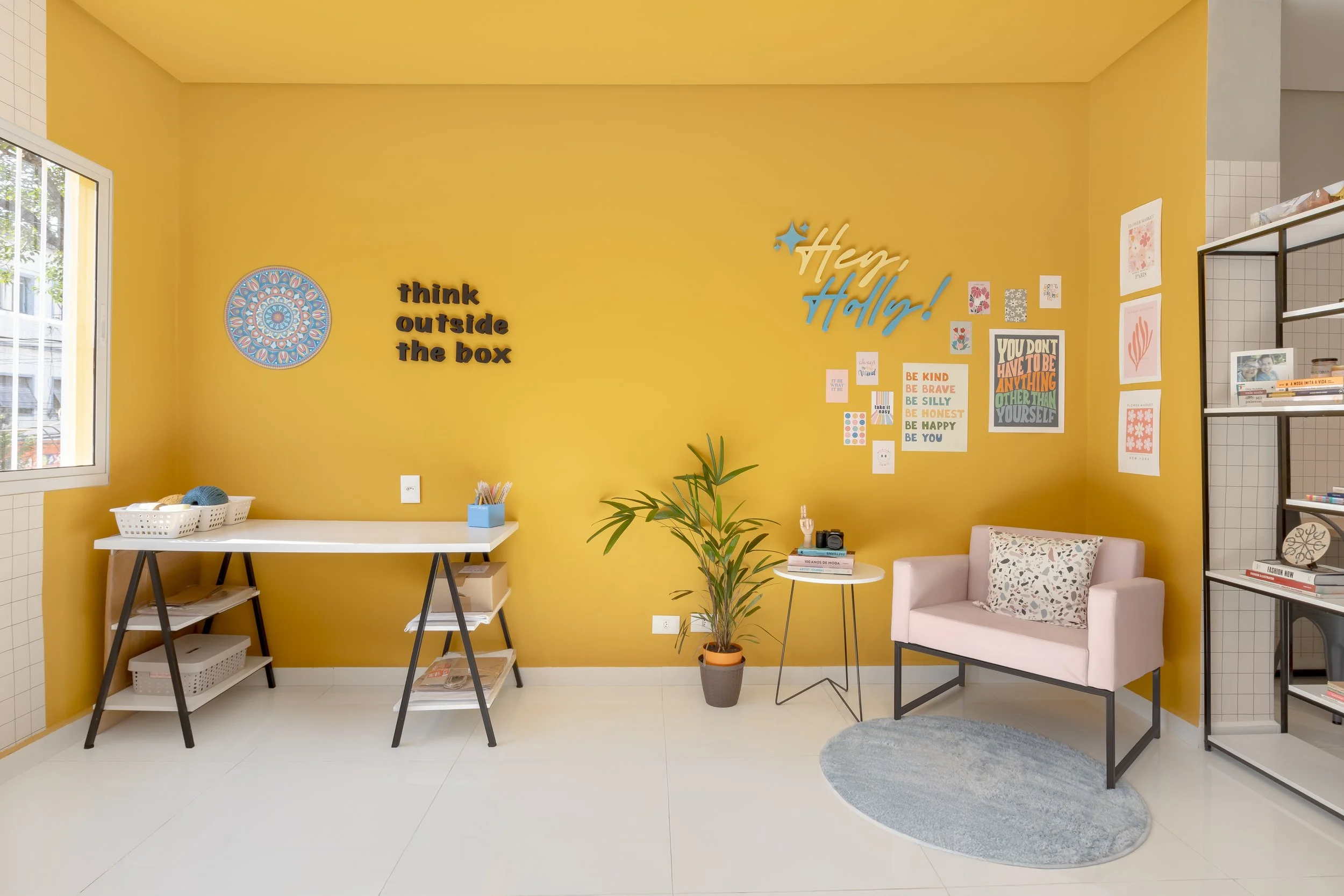 Sala de estar com parede amarela, mesa branca, prateleira, vaso com planta, sofá rosa claro, almofada decorativa, prateleira de livros, cartazes motivacionais na parede e gaveta com decoração.