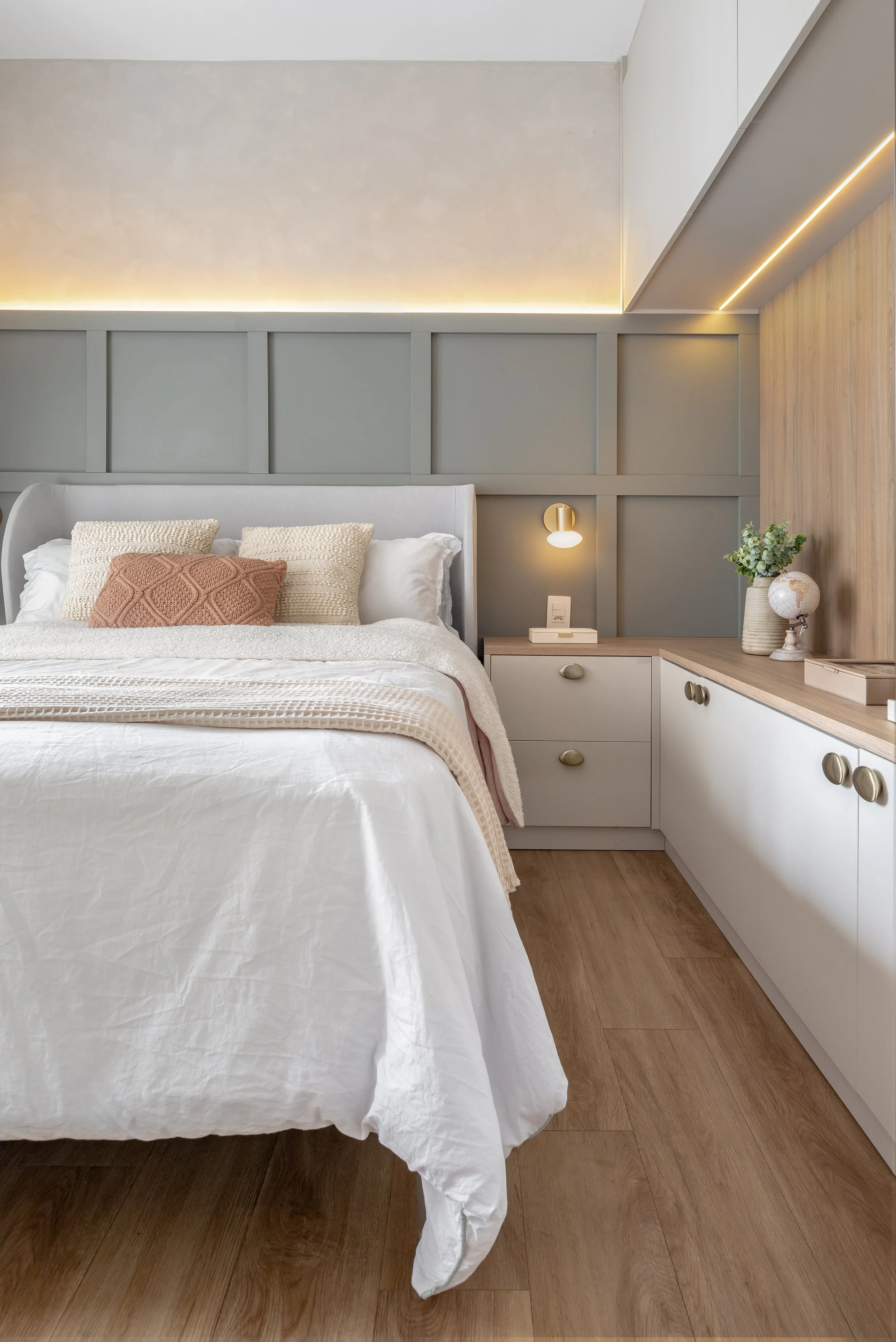 Cama de casal com cabeceira acolchoada, almofadas decorativas, roupa de cama branca, lado de um criado-mudo com uma luminária, plantas e objetos decorativos, parede com painel de madeira e iluminação indireta.