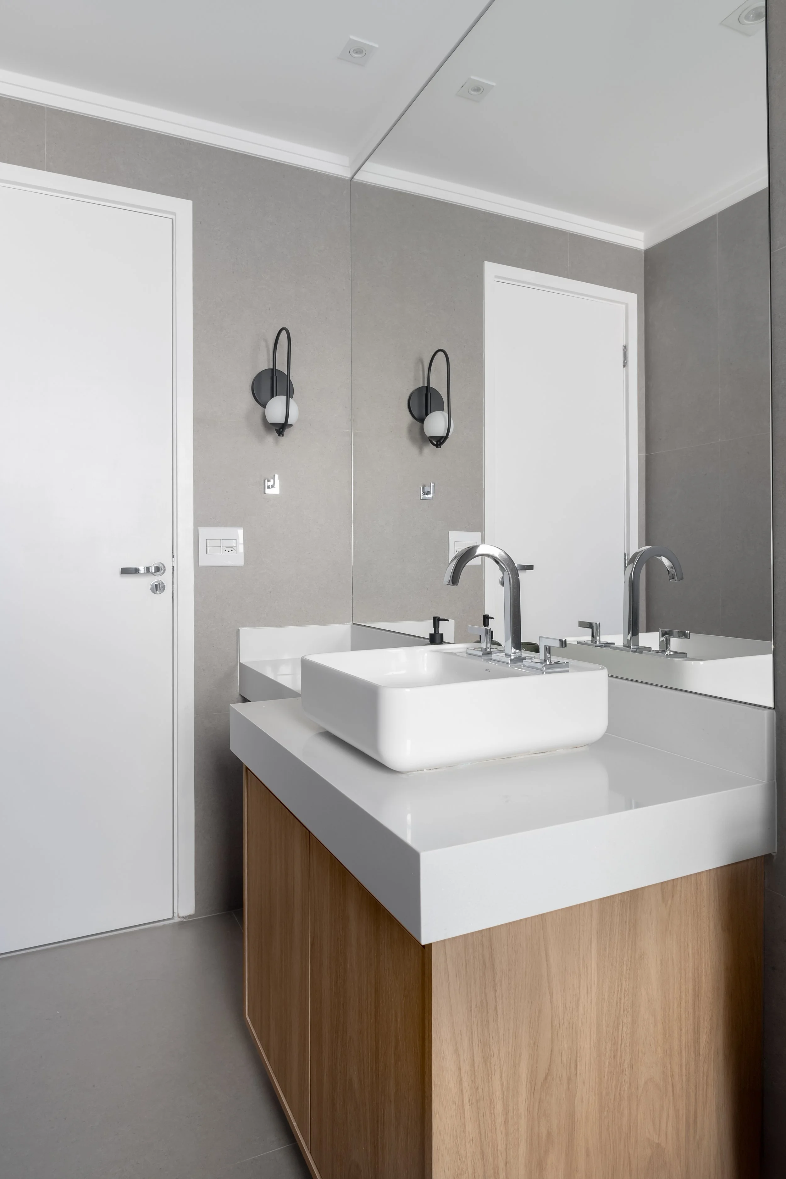 Espelho grande refletindo uma pia com cuba branca sobre bancada de madeira e parede cinza, em um banheiro moderno.