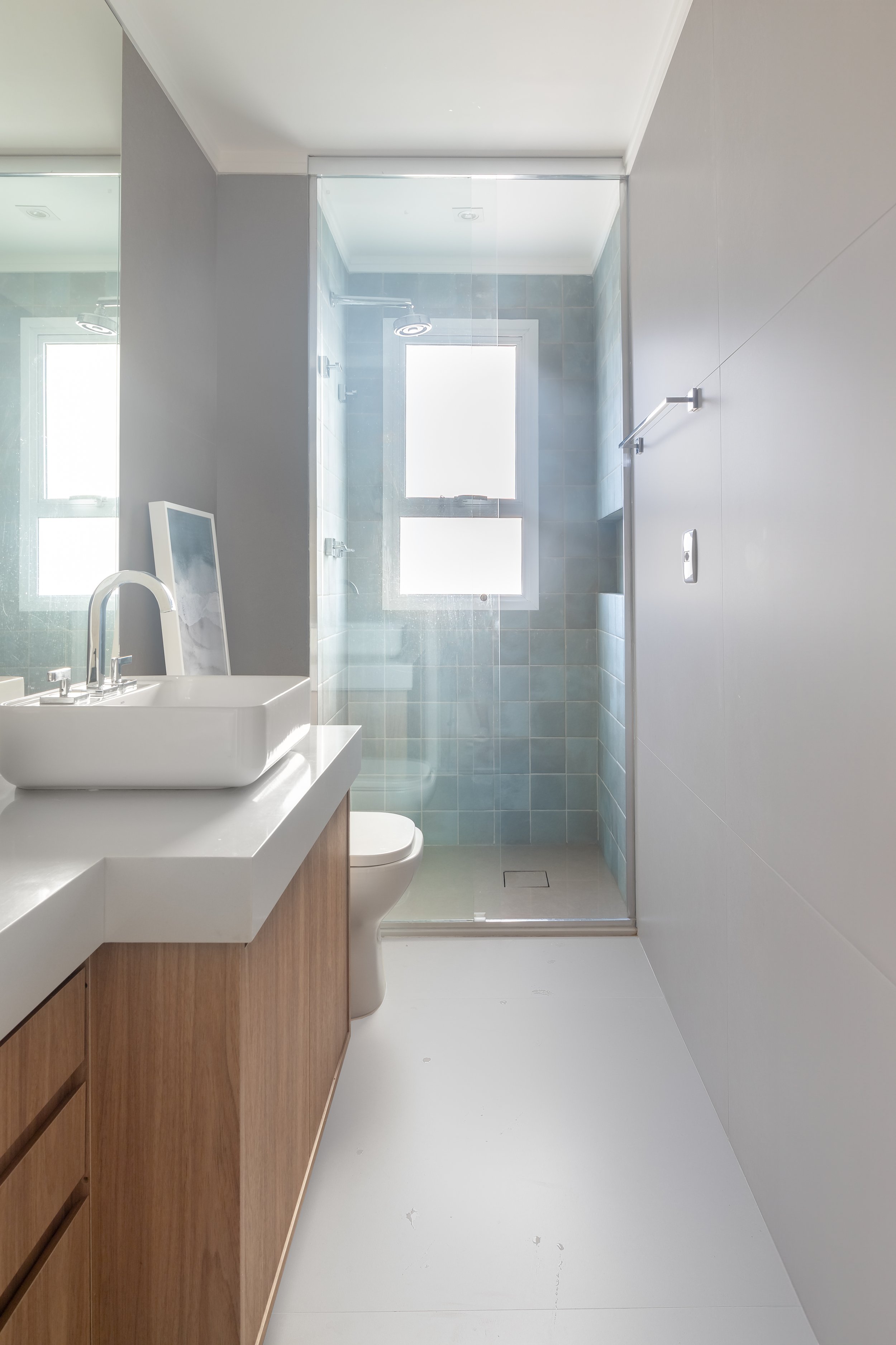 Banheiro moderno com pia de madeira, vaso, box de vidro com janela na ducha e paredes claras.