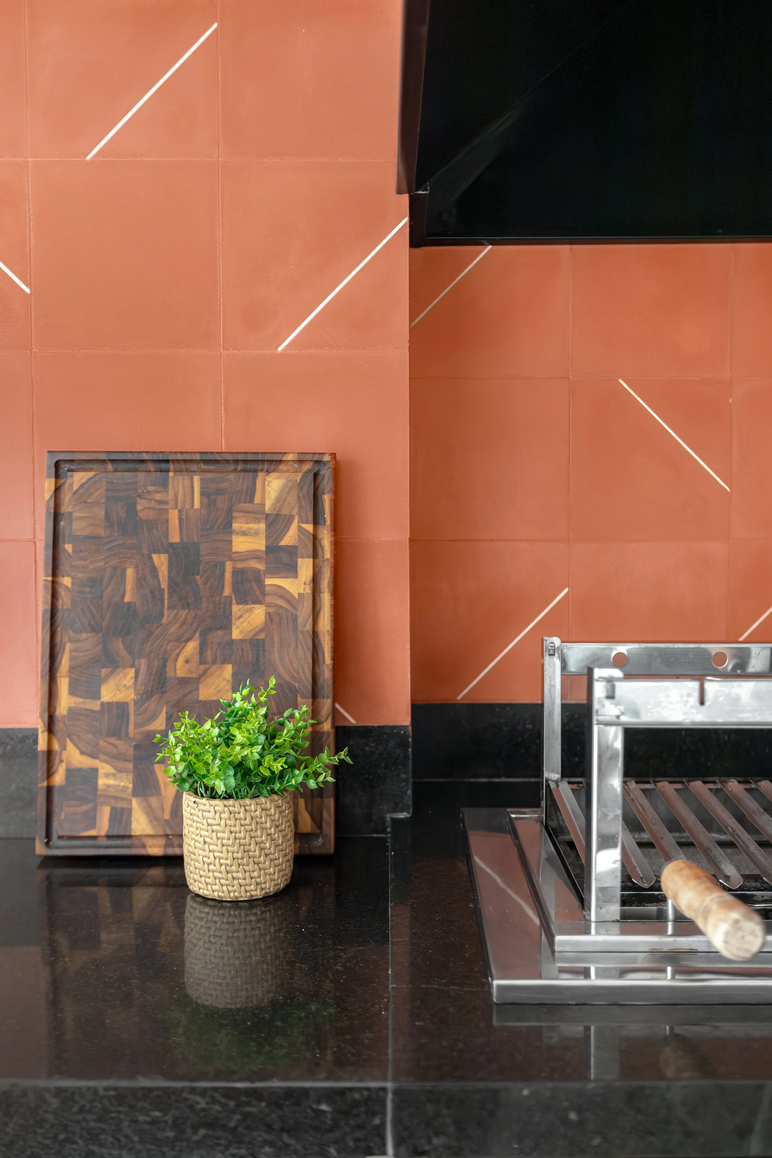Decorativo espaço com piso de azulejo vermelho, uma saia de madeira com padrão geométrico, uma planta verde em vaso de crochê, uma estrutura de metal inoxidável, uma parede de azulejo vermelho com linhas brancas, uma folha de papel alumínio, uma luz 