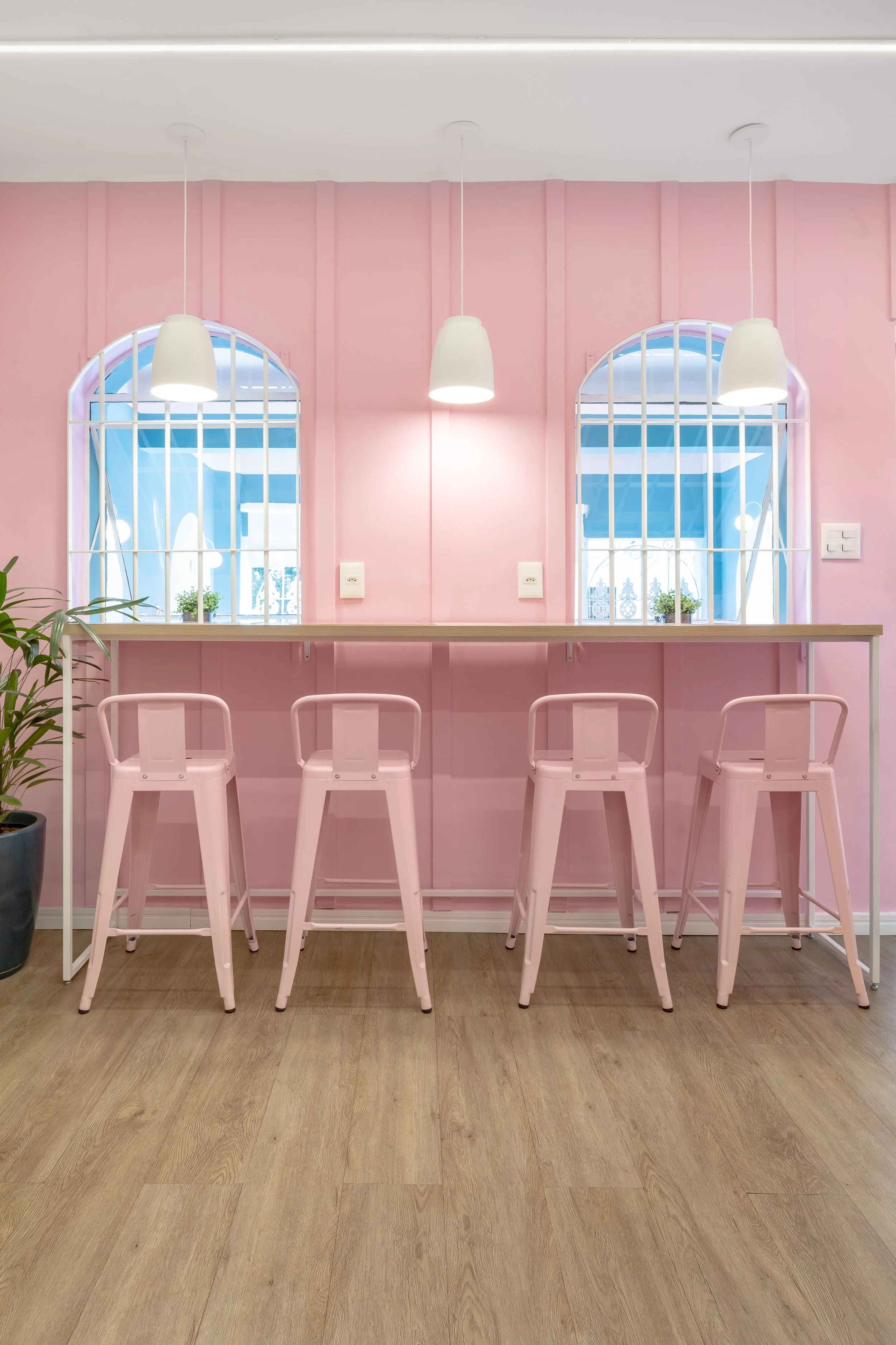 Barra com quatro cadeiras de ferro pintadas de rosa em frente a paredes rosa claro, com duas janelas arqueadas com grades brancas e plantas ao lado.