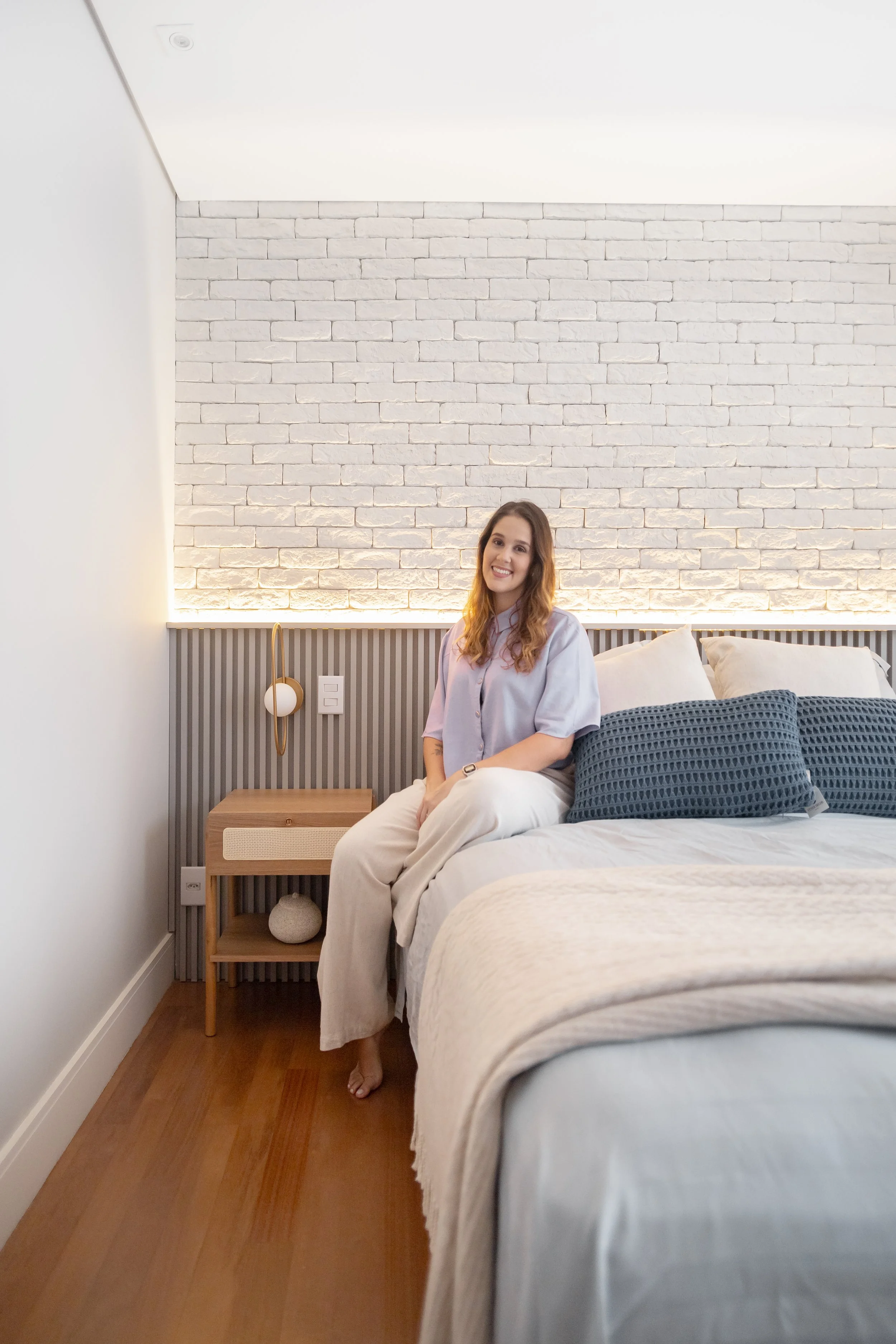 Mulher sorridente sentada na cama de um quarto decorado com parede de tijolos brancos, cabeceira de painéis ripados e luminária de parede.