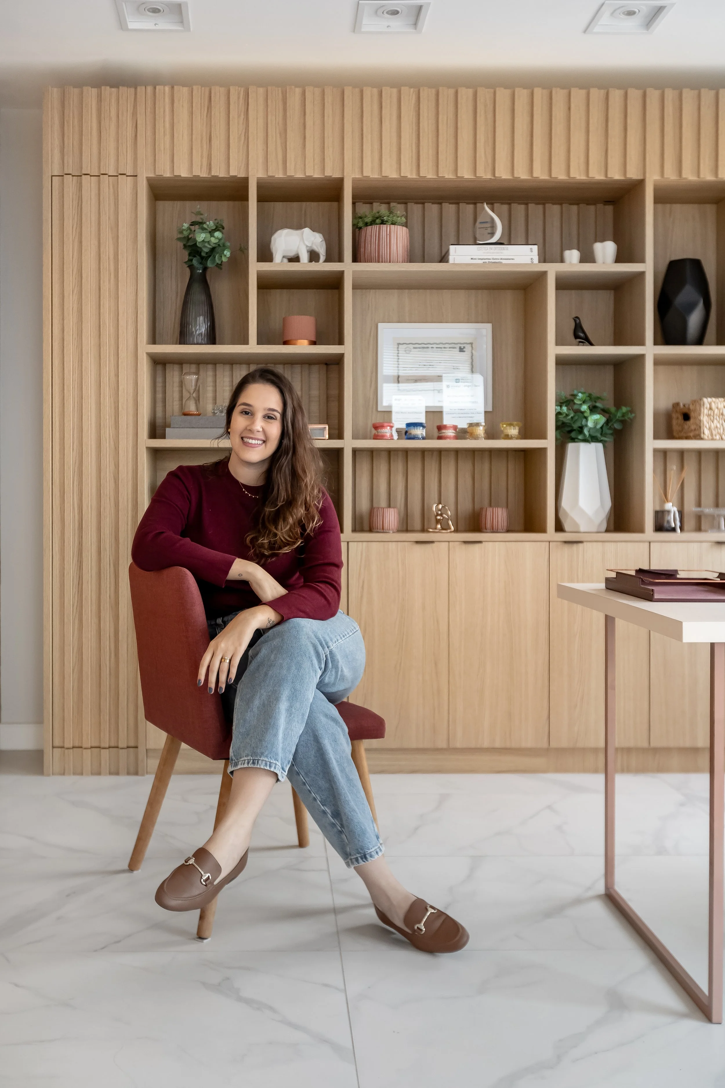 Mulher sorridente sentada em uma cadeira vermelha, com roupas casuais, em um ambiente com parede de madeira decorada com prateleiras que exibem vasos, livros, objetos decorativos e uma planta.