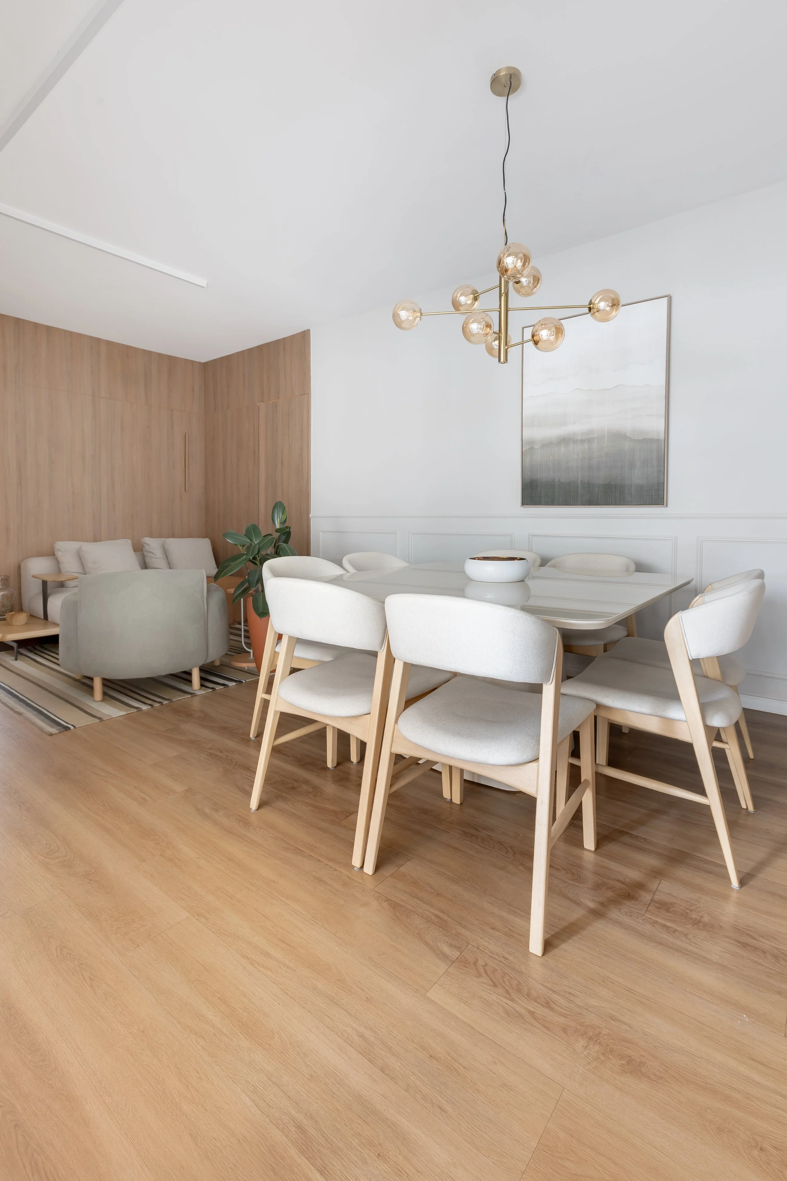 Sala de jantar com mesa branca, cadeiras brancas com pés de madeira, luminária moderna com lâmpadas de vidro e quadro com paisagem na parede.