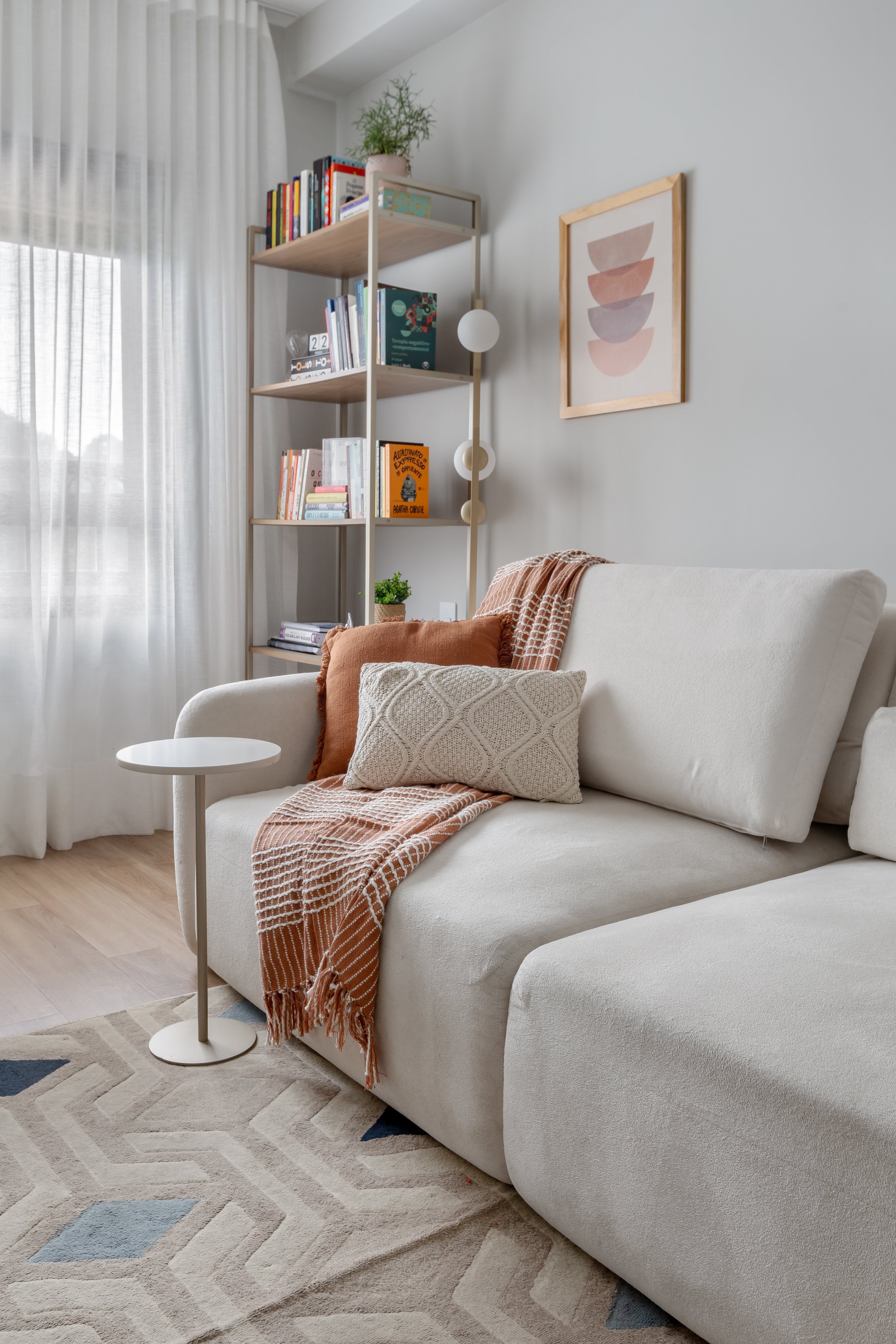 Sala de estar com sofá bege, almofada branca com detalhes em lã, manta listrada em tons de rosa e laranja, tapete geométrico com cores neutras e detalhes em azul. Há uma parede com quadro de formas abstratas em tons de rosa, lavanda e bege. Uma estan