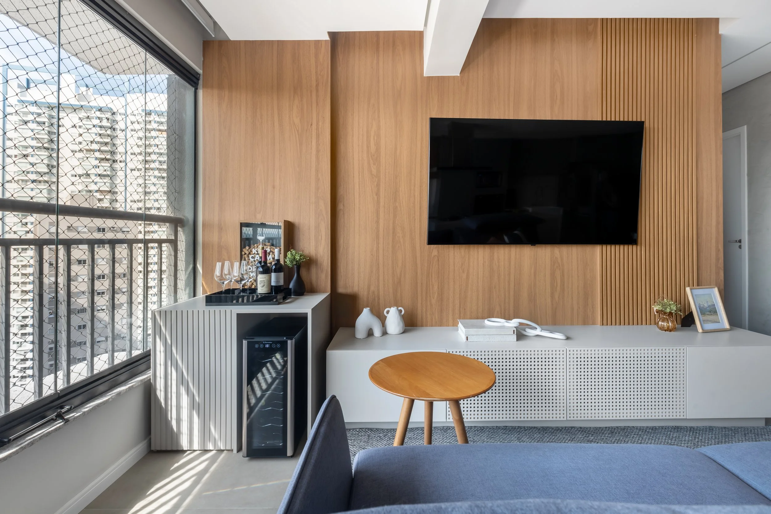Sala de estar com parede de madeira, TV de tela plana, balcão branco com decoração, vinho, copos e uma mini geladeira, ao lado de uma janela extensa com grades, um pequeno banco de madeira, uma planta, uma moldura de quadro e um sofá cinza.